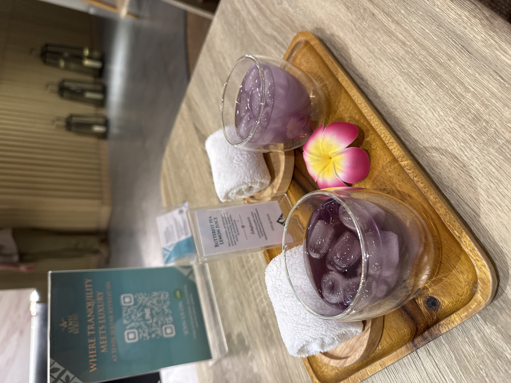 Captured in what appears to be a high-end spa or resort in Bangkok, Thailand, the image presents a close-up, overhead shot of a wooden tray arrangement. Two glasses filled with a purple liquid, likely a Butterfly Pea Lemon Juice, are the focal points. Each glass sits on a small, round wooden coaster, cushioned by a rolled white towel. A pink and yellow flower adds a touch of color to the display. Positioned beside the drinks are two promotional cards. The first card, titled "Butterfly Pea Lemon Juice" in English and Thai, provides a brief description. The second card, predominantly teal, features a QR code and the phrase "WHERE TRANQUILITY MEETS LUXURY" along with "SENSE SERENE RETREAT" indicating the location. The tray rests on a light-colored, wood-grain surface. In the blurred background, there's a glimpse of a hallway with decorative elements along the wall, suggesting the interior design of a luxurious spa environment.