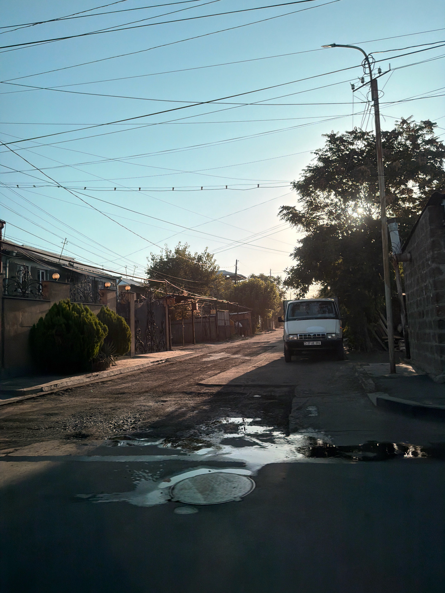 The image depicts a street in Yerevan, Armenia, bathed in the warm light of a setting sun. Overhead, a tangle of electrical wires crisscrosses the clear blue sky, a common sight in urban Armenia. A streetlamp on a utility pole stands to the right, its light fixture angled downwards.
The street itself is paved with asphalt, showing signs of wear and tear, with puddles of water reflecting the sky and surrounding elements. A manhole cover is visible in the foreground, partially submerged in a puddle.
On the right side of the street, a white van is parked, its headlights off. A license plate is visible, reading "37 UE 396". Behind the van, a dense tree with sunlight filtering through its leaves creates a dappled effect. Further back, a brick wall forms the side of a building.
To the left of the street, a sidewalk runs alongside a row of houses. The houses have stone facades and ornate wrought-iron gates and fencing. Two evergreen bushes are neatly trimmed in front of one of the properties. Another house further down the street has a corrugated metal roof. The overall impression is of a residential street in a city transitioning from day to evening.