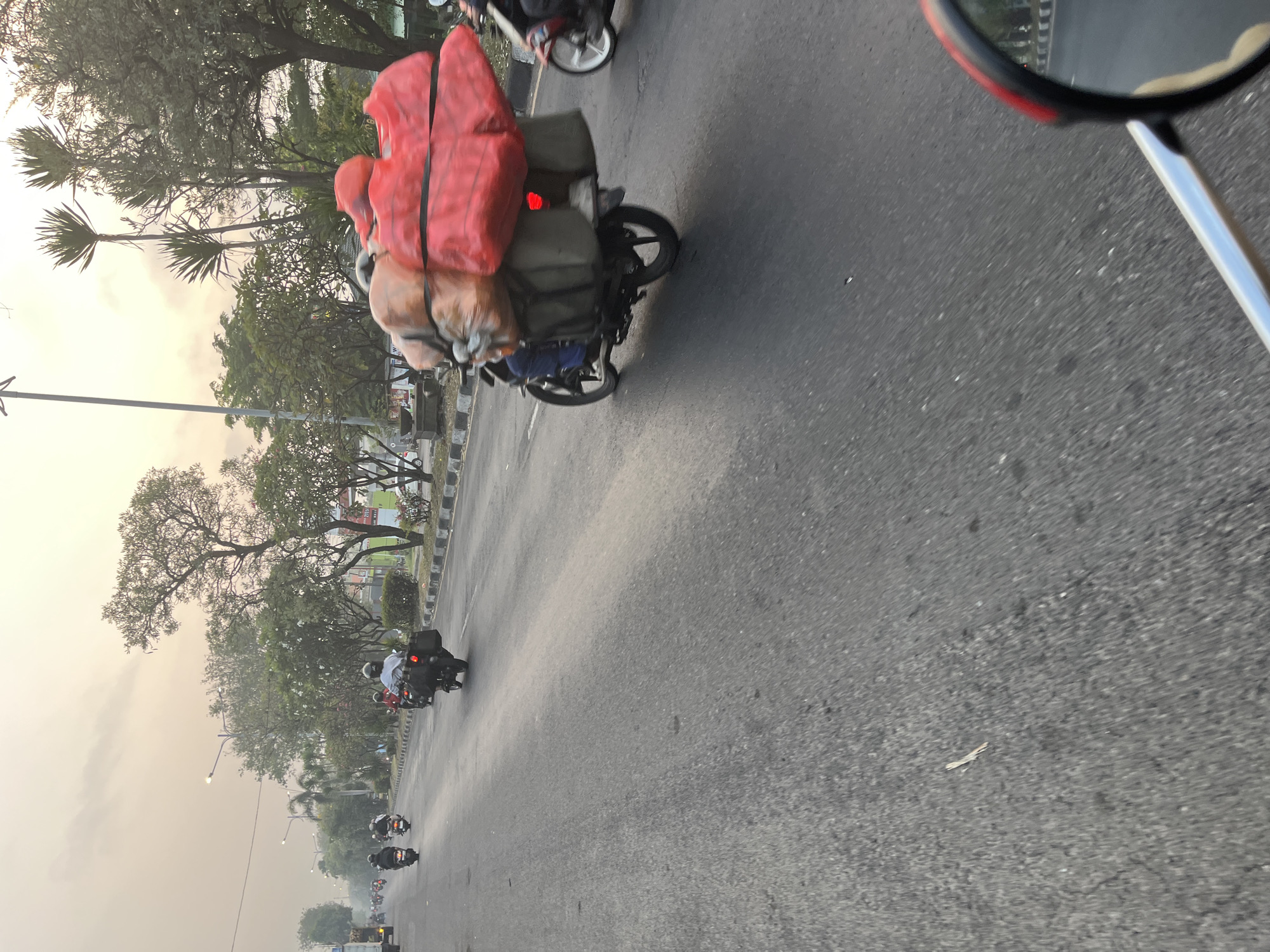 In Kludan, Indonesia, at dusk, a motorcycle transports an oversized load of red and brown bags, secured with black straps, down a paved road. The motorcycle has a black storage box and a red taillight. The rider is wearing a white shirt and a helmet. Several other motorcycles are also on the road, some carrying passengers. The scene is lined with trees and a sidewalk. The sky is a pale orange, indicating the end of the day. The perspective suggests the image was taken from another motorcycle.