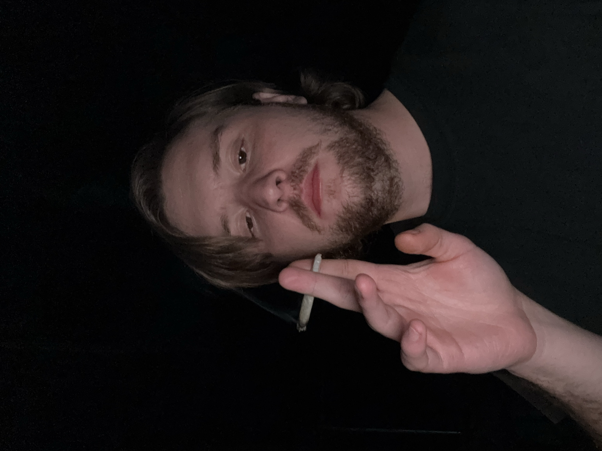 A man with light hair and a beard is holding a lit cigarette in his right hand. He is wearing a black shirt and appears to be indoors, given the dark background. The location is not identifiable as being in Cañada de Madero, Mexico, based on the visual information provided.