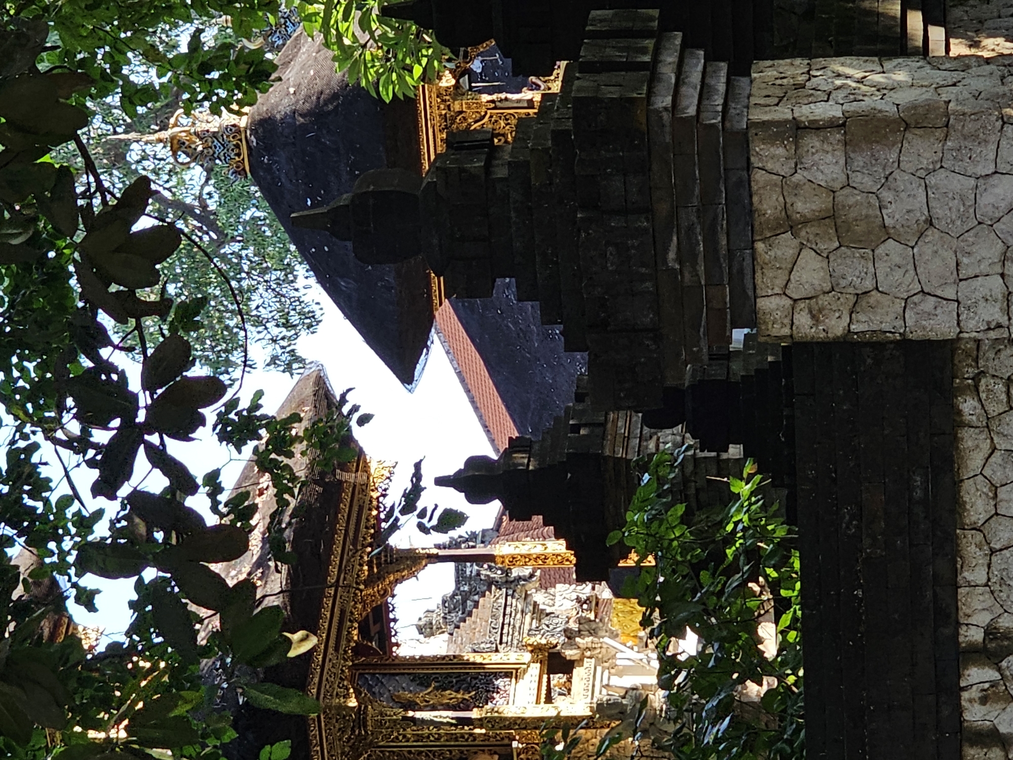 The image depicts a section of a temple complex, likely in Denpasar, Indonesia, given the architectural style. The foreground is dominated by a stone wall constructed from irregular, light-colored stones, stacked in a mosaic-like pattern. Above this, dark grey, rectangular stones form a more structured wall. Emerging from behind this wall are tiered stone structures with pointed finials, characteristic of Balinese temple architecture.
To the left and right, glimpses of ornate temple structures are visible. The structure on the left features intricate gold detailing on its roof and facade, with vertical decorative elements and what appears to be carved panels. The roof of this structure is dark, possibly thatched or tiled. Behind the tiered stone structures, a dark, multi-tiered roof is visible, topped with a golden, elaborate spire or ornament. This roof also appears dark, possibly covered in traditional materials.
Green foliage from trees overhangs the scene from the top and the middle ground, casting shadows and partially obscuring the view of the sky. The lighting suggests it is daytime, with sunlight filtering through the leaves and illuminating parts of the golden decorations. No people are visible in the image. The overall impression is one of a serene, traditional temple setting within a lush environment.