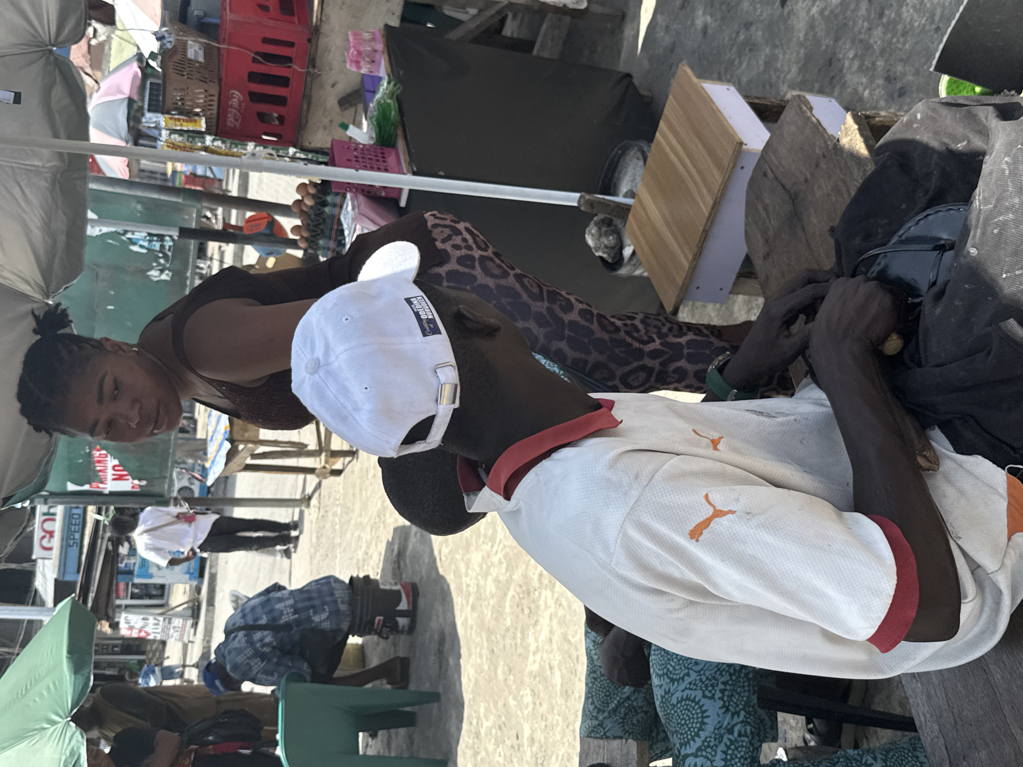 In Ado, Nigeria, a woman is looking at a man who appears to be working on or repairing a black bag or item. The man is wearing a white shirt with orange accents and a white cap with a logo on the back. He is sitting on a wooden structure. The woman is wearing a brown top and printed leggings. They are under an umbrella, possibly a market setting, with various objects like crates, boxes, and other people visible in the background. There are commercial signs visible in the background as well.