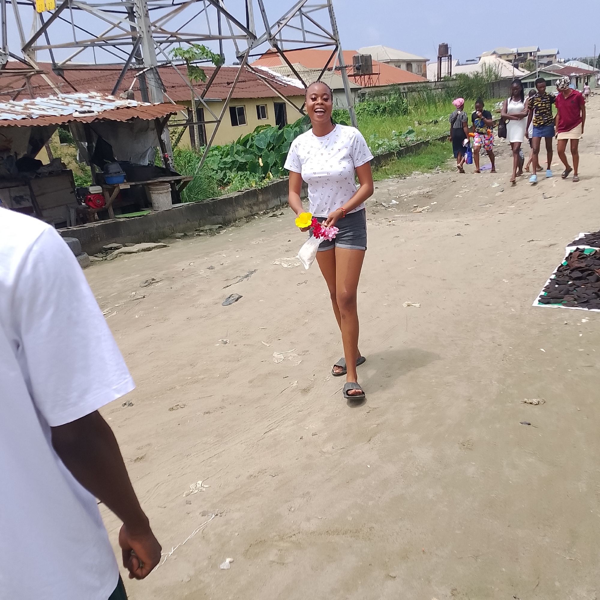 The image shows a group of people on a dirt road in Ado, Nigeria. A young woman in the foreground is smiling and holding two flowers. She is wearing a white t-shirt with a patterned print and dark shorts. Behind her, several other people are walking along the road. To the left, there is a makeshift shelter with a rusted tin roof, and in the background, there are houses and a tall metal structure, possibly a transmission tower. The ground is sandy and littered with debris. The scene appears to be a casual outdoor gathering or a moment in daily life in a Nigerian city.
