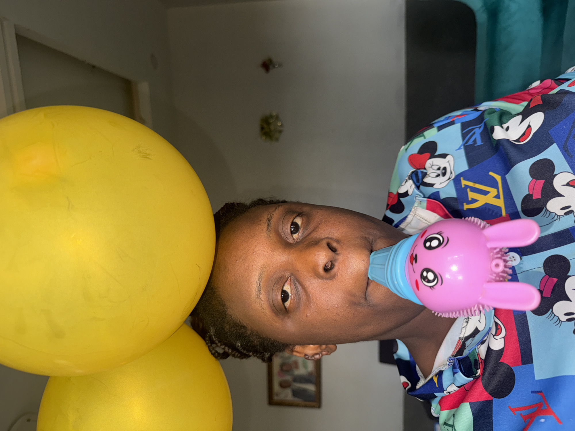 A dark-skinned person is holding a pink toy bunny-shaped bottle to their mouth. Two yellow balloons are to their left. The person is wearing a light blue shirt with cartoon characters. An interior background is visible behind them. There is no verifiable evidence to support the location being Sabon Tasha, Nigeria.