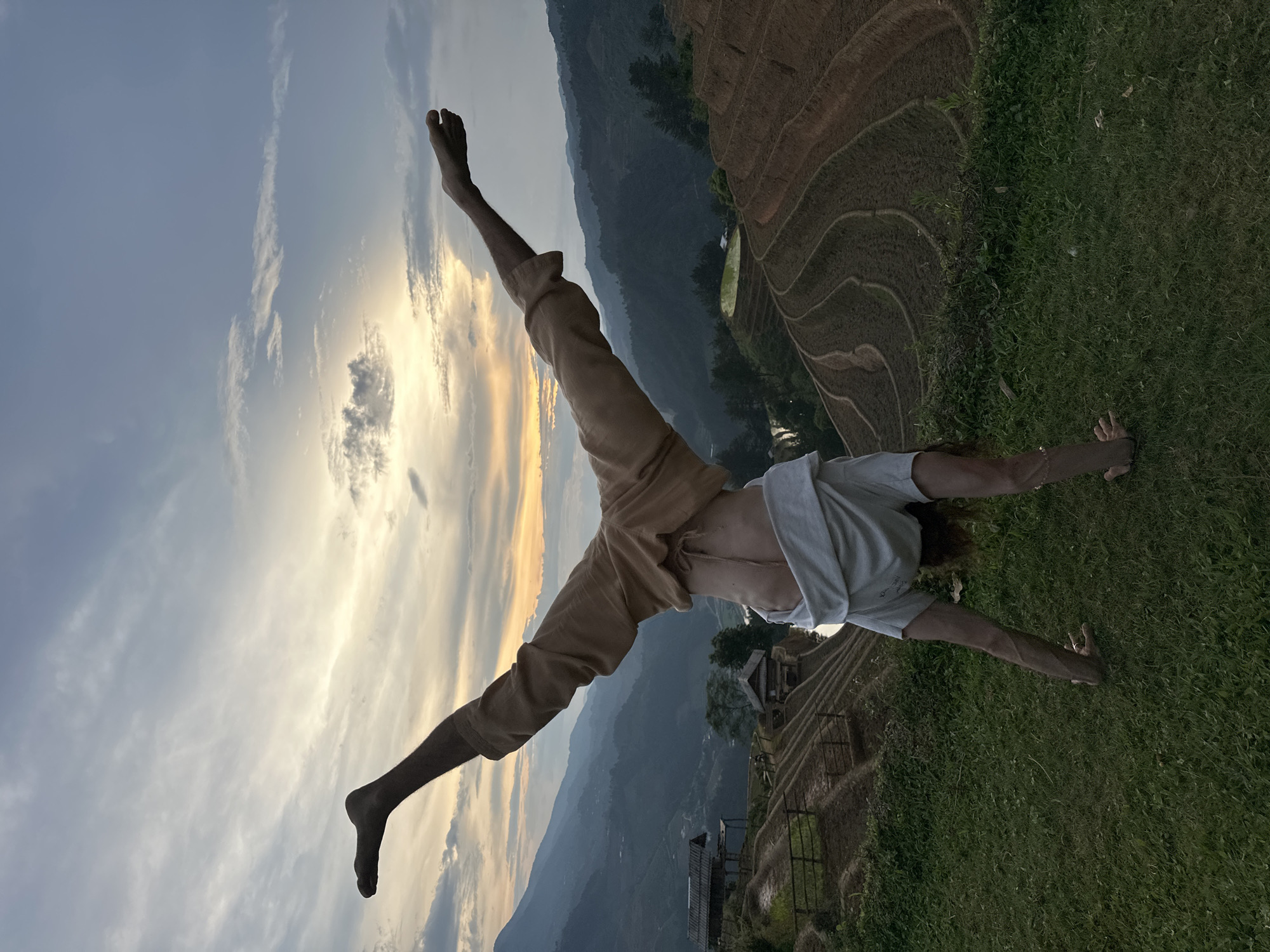 This image, rotated 90 degrees clockwise from its natural orientation, depicts a person performing an acrobatic pose against a magnificent landscape identified as Mù Cang Chải, Vietnam. The individual is positioned in a dynamic, inverted 'V' shape, suggesting a handstand or cartwheel. Their bare feet are extended upwards towards the left of the frame, silhouetted against the sky, while their hands support their weight on a patch of green grass in the lower right. They wear light-colored trousers and a light-toned t-shirt.
The background unfolds into an expansive vista of meticulously carved rice terraces, a hallmark of Vietnam's mountainous agricultural regions. These terraces form intricate patterns across rolling hills, with distant mountains fading into a soft haze. Several small, traditional buildings are visible nestled within the terraced landscape.
The scene is captured during what appears to be sunset or sunrise, evidenced by the warm, golden light emanating from the horizon on the left. This illumination casts a radiant glow on scattered clouds, creating a vibrant contrast with the softer blue of the sky and the darker terrain. The dramatic lighting emphasizes the person's form against the natural splendor. The composition conveys a sense of freedom, strength, and harmony between the human subject and the vast, beautiful environment. No visible text is present in the image.