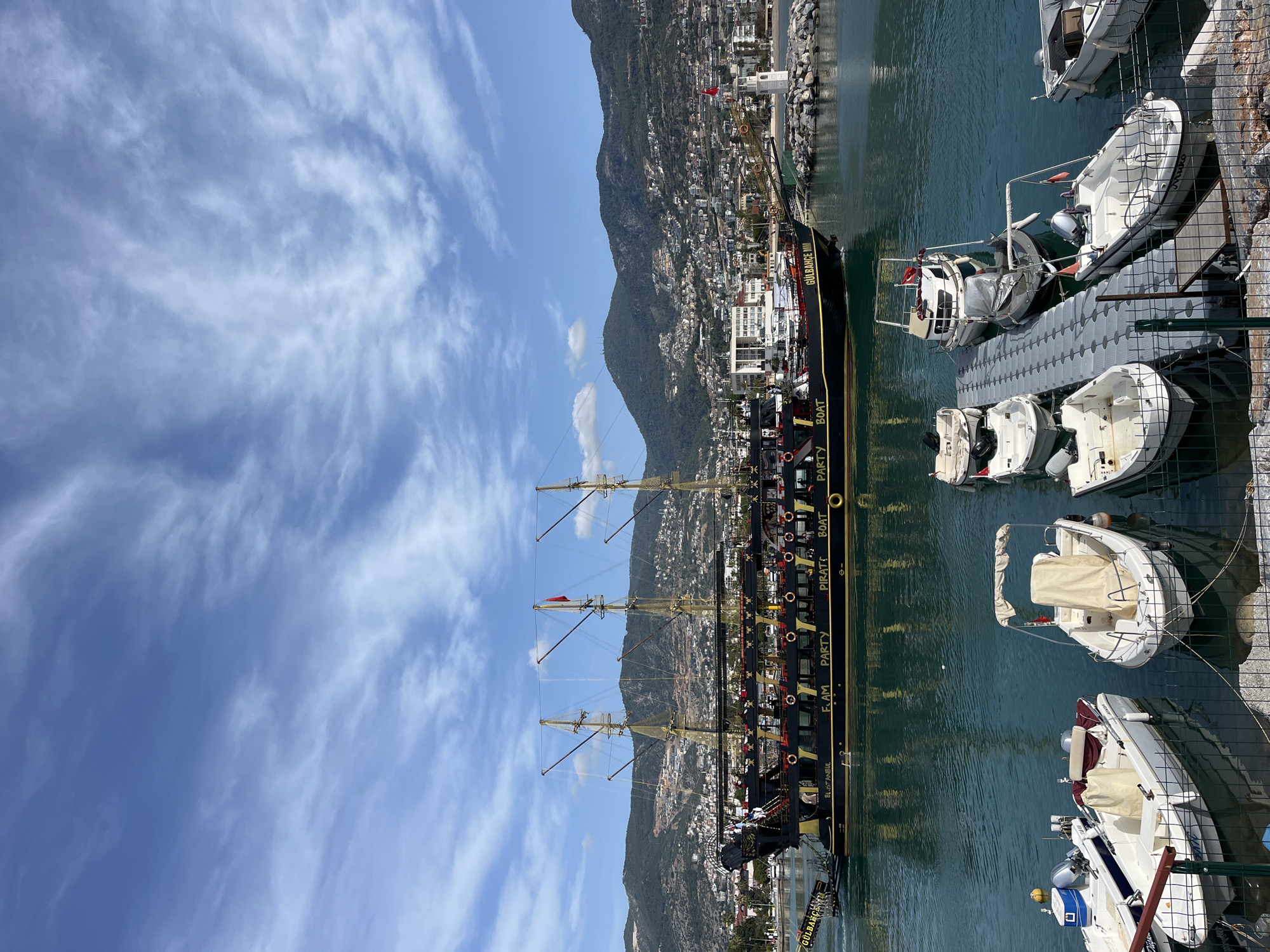 The image depicts a harbor scene in Alanya, Turkey. A tall, black pirate-themed party boat named "GULBAHCE IV," is docked alongside a gray floating dock. Several small, white boats are moored alongside the dock. A mountain range serves as the backdrop to the city, which is built along the shoreline. The sky is a bright blue with light, wispy clouds. A metal fence is partially visible in the foreground, near the docked boats. The water in the harbor is a dark green color. There are no visible people in the image.