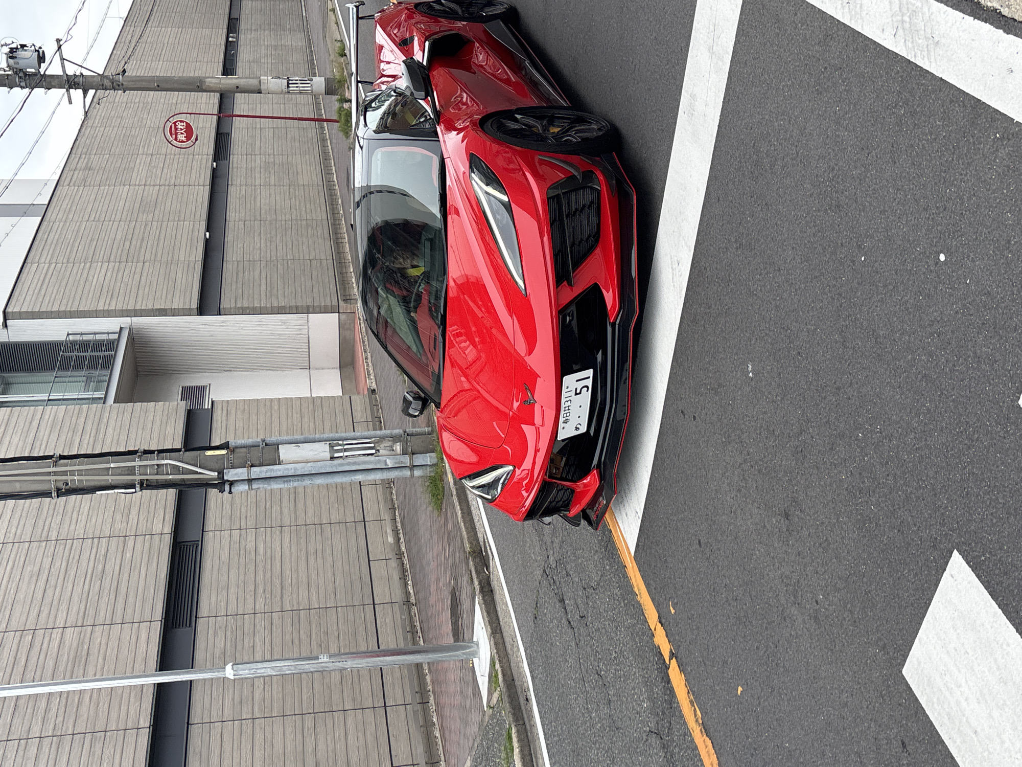 A bright red Chevrolet Corvette C8 is parked on a street in Nagoya, Japan. The car is angled towards the viewer, with its front end slightly to the left. The license plate is visible and reads "名古屋311 51". The car has a black roof and black accents, including a prominent rear spoiler. It is positioned on asphalt pavement marked with white pedestrian crossing lines and a yellow line. In the background, a modern building with horizontal wooden-like siding is visible. A utility pole stands to the right of the car, and a red and white fire alarm sign is mounted on it. The sky is overcast. The scene appears to be a typical urban street in Japan.