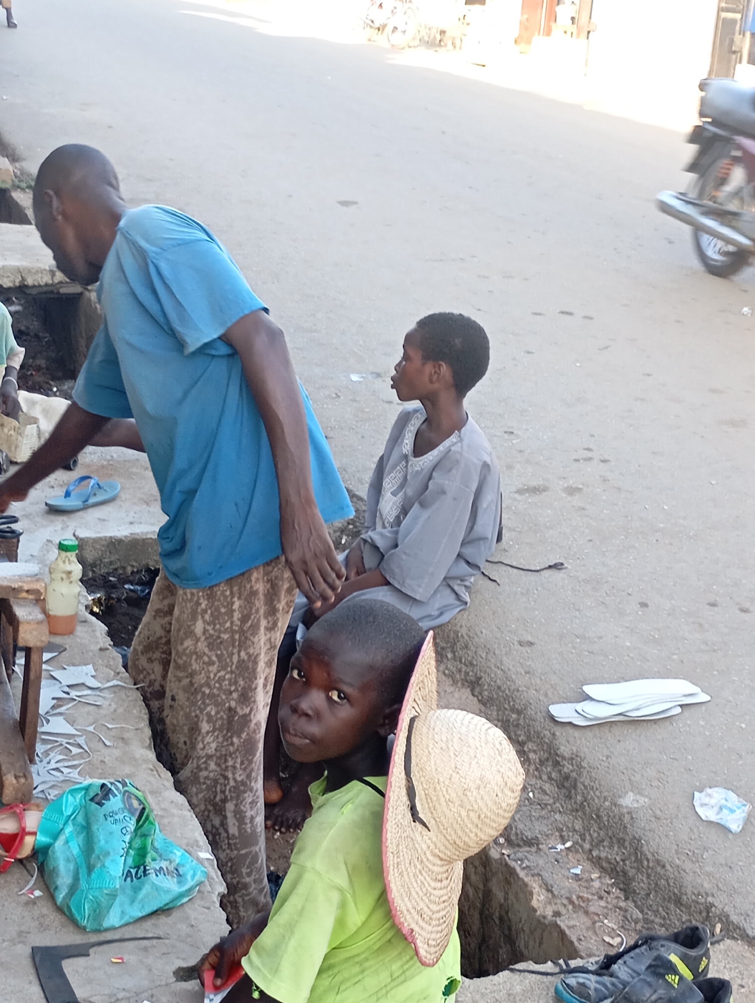 The image depicts a street scene in Shendam, Nigeria, focusing on individuals engaged in what appears to be a shoe-repair or craft activity. In the foreground, a young boy wearing a lime green t-shirt and a straw hat is looking towards the camera. To his left, an adult male in a blue t-shirt and patterned trousers is bending over, seemingly working on something on a raised concrete surface. Another young person in a light gray garment sits to the right of the man, observing. The environment includes a dusty road surface, a concrete curb, and various objects related to the activity, such as cut pieces of material resembling leather or rubber, a plastic bag, a bottle with a yellowish liquid, and a pair of black athletic shoes. The overall impression is of a community setting where informal trade or repair work is taking place outdoors.
