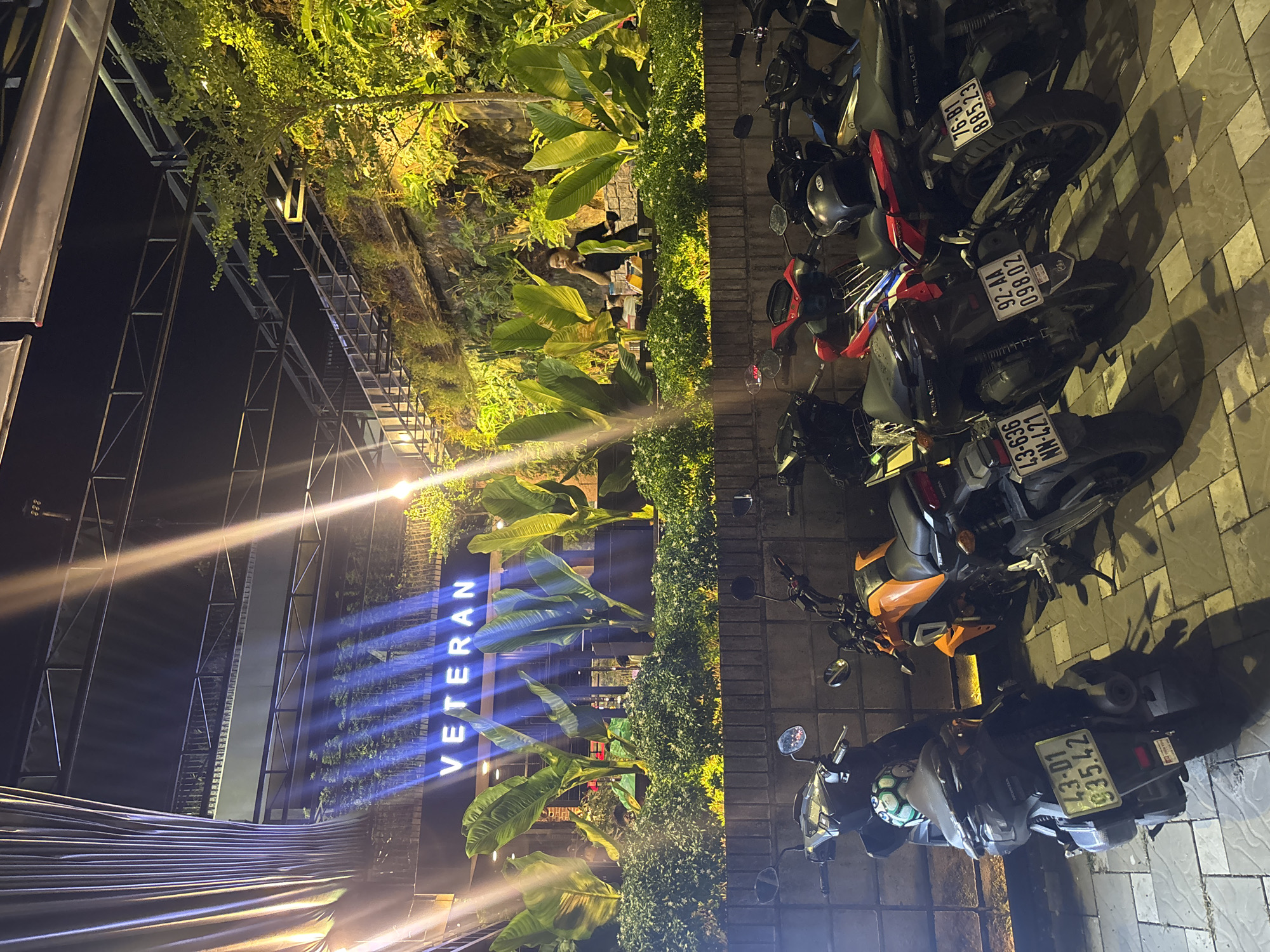 In Đà Nẵng, Vietnam, at night, a neon sign spelling "VETERAN" illuminates a building facade. Large green banana leaves and lush green bushes are prominently displayed in front of the building. In the foreground, several parked motorcycles are visible. One motorcycle is black with a license plate reading "43-D1 935.42". Next to it, an orange motorcycle has a license plate "43-636 NN-421". Further down, a red and black motorcycle has a license plate "92-AA 098.02", and another motorcycle behind it has a plate "76-B1 885.23". A person is seated at a table behind the plants, appearing to eat. The scene suggests an outdoor seating area of a restaurant or establishment in the city.
