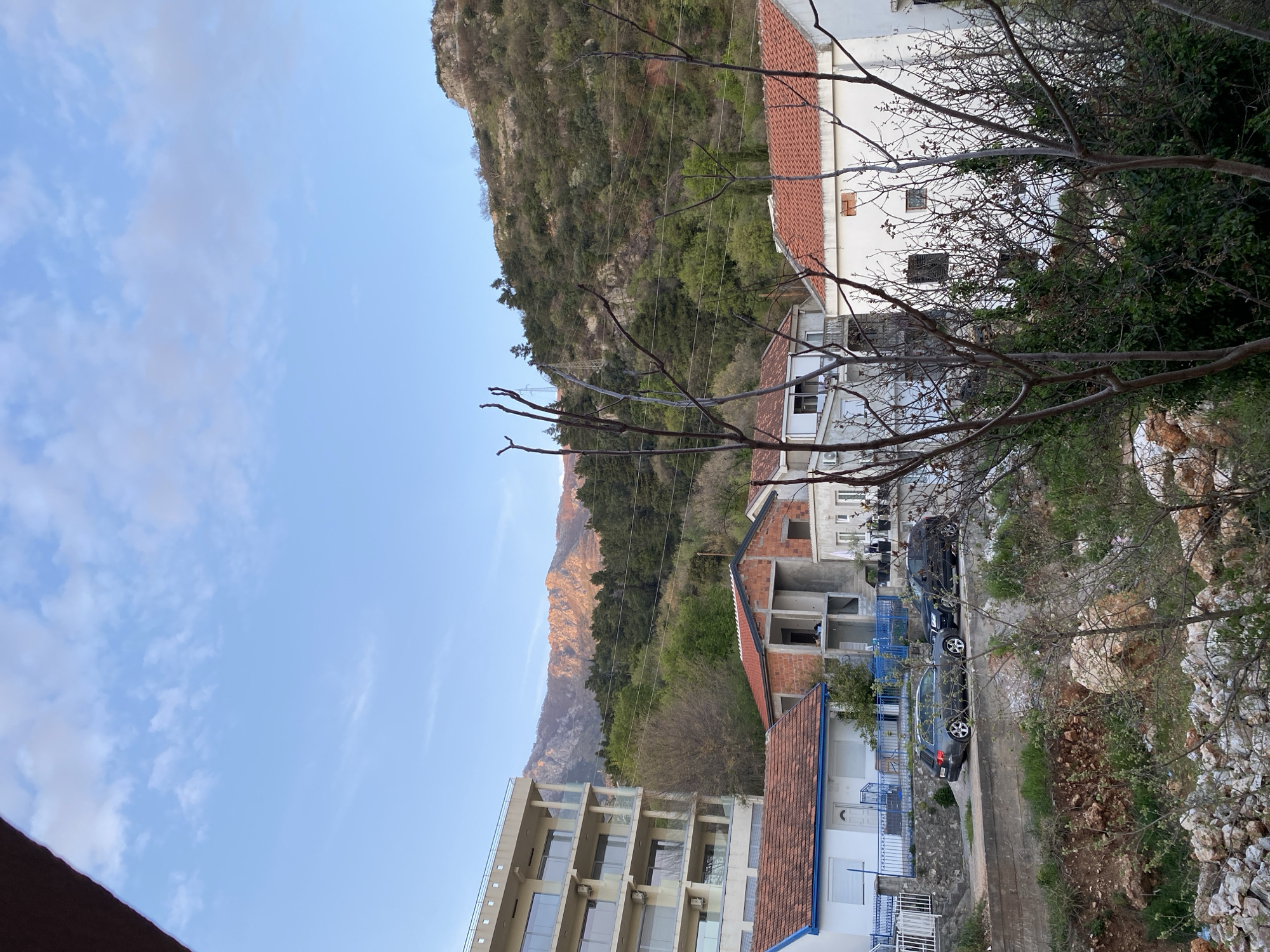 The image depicts a street scene in Budva, Montenegro, featuring a mix of residential buildings and natural elements. The buildings are a combination of older structures with red tile roofs and a modern multi-story building with glass balconies. A building under construction is visible. Several cars are parked along the street. A rugged mountain face rises in the background, partially illuminated by the setting sun. Bare tree branches are prominent in the foreground, and there is some vegetation along the roadside. The sky is blue with scattered clouds.