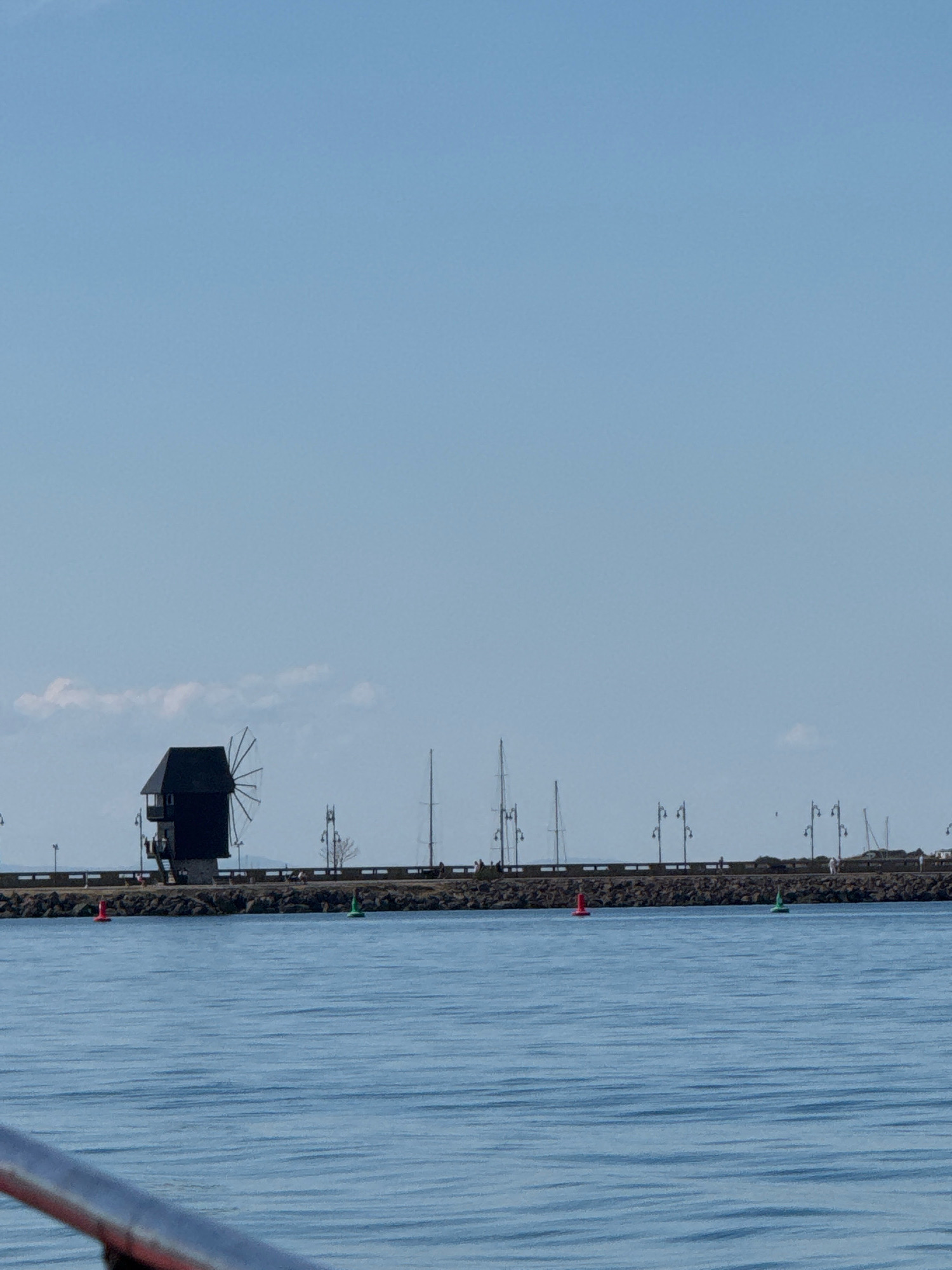 The image depicts a view of the Nesebar Peninsula in Nesebar, Bulgaria, from the water. In the foreground, the calm, blue water reflects the clear, pale blue sky. A dark, traditional windmill, a prominent landmark, stands on a stone embankment along the coast. The embankment is lined with streetlights and a low wall. Beyond the windmill, several sailboats and masts are visible. Two buoys, one red and one green, float in the water near the embankment. A few people are seen walking along the embankment. The overall scene is tranquil and depicts a sunny day.