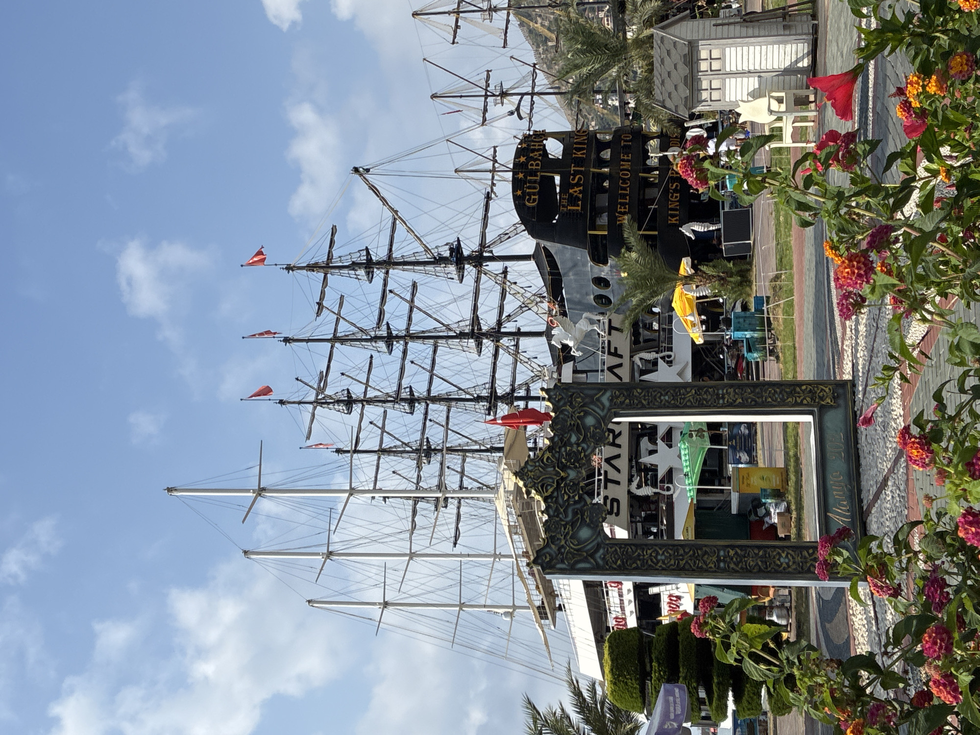 The image depicts a large, multi-masted sailing ship docked in a harbor, likely in Alanya, Turkey, given the inscription "Alanya 2023" on the ornate frame in the foreground. The ship is named "GULBAHCE THE LAST KING" and has the text "WELCOME TO KING'S PARADISE" written on its stern. Several red flags are visible on the ship's masts. In the foreground, a decorative, baroque-style frame stands in front of blooming flowers, with a seating area and a small shed visible behind it. Palm trees and other greenery are scattered throughout the scene. The sky is blue with scattered clouds, suggesting a daytime, possibly sunny, weather condition. The overall scene appears to be a tourist attraction or a popular waterfront area.