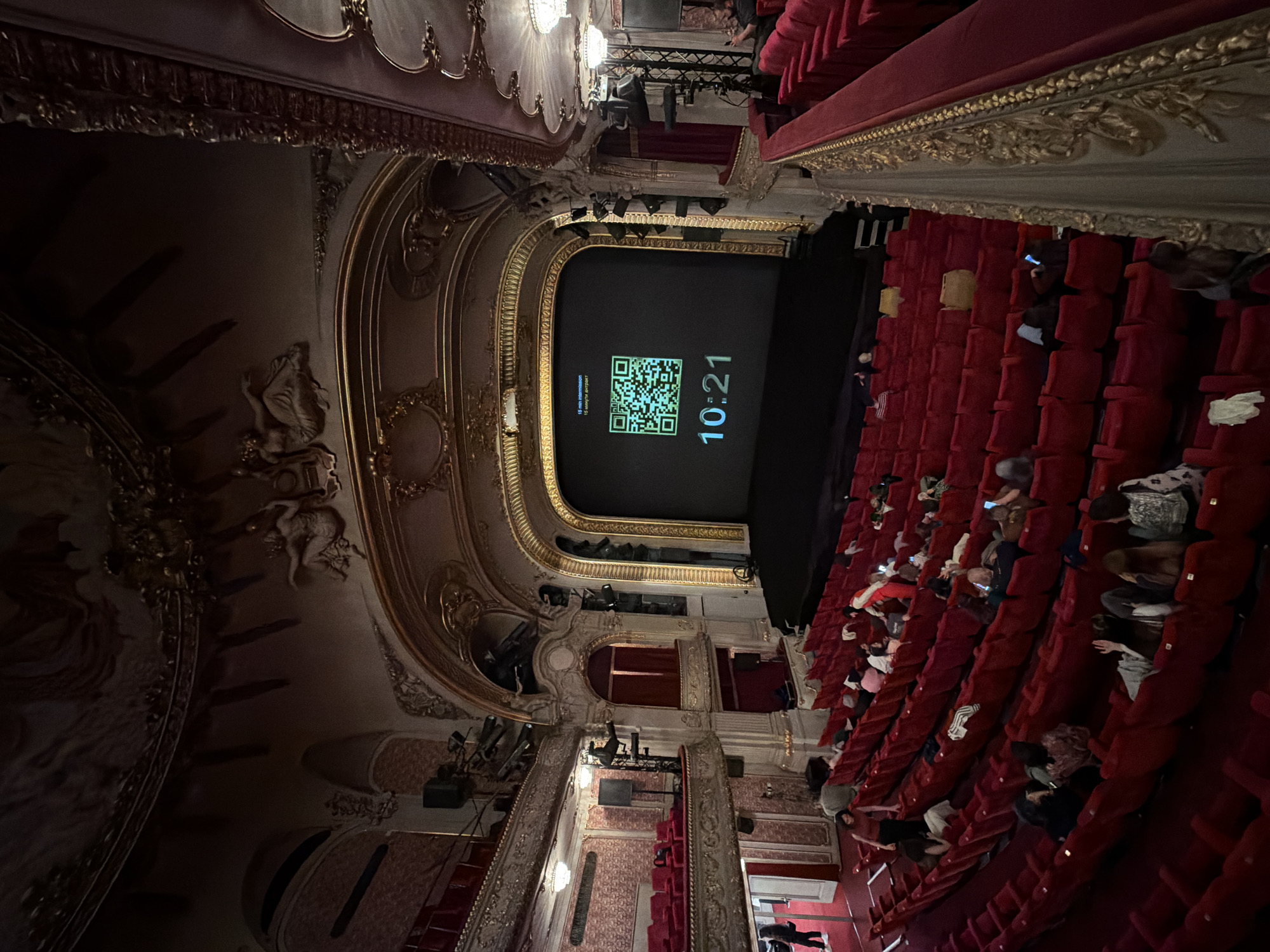 This image presents an angled, overhead view inside a grand, ornate theater or opera house, likely in Sofia, Bulgaria, given the context. The interior showcases opulent classical architecture with intricate gold leaf detailing and decorative stucco work adorning the proscenium arch, balconies, and upper walls.
The auditorium is filled with rows of plush red velvet seats, where a moderate number of audience members are scattered, appearing to be settled and waiting. Some individuals are seen looking at their mobile phones.
The focal point is the stage, where a large black screen is illuminated. It displays a prominent green QR code on the left side, accompanied by text. Below the QR code, the text reads "15 min intermission" and "15 минути до края" (Bulgarian for "15 minutes until the end" or "15 minutes until the break"). To the right of the QR code, a digital clock shows the time "10:21". This setup indicates either the start of an intermission countdown or a pre-show waiting period.
The lighting is dim, characteristic of an indoor performance venue before a show or during a break, with the screen being the brightest light source. Several spotlights and stage lighting fixtures are visible above the stage and along the balcony edges. The overall atmosphere is one of calm anticipation within a richly decorated, historical setting.
