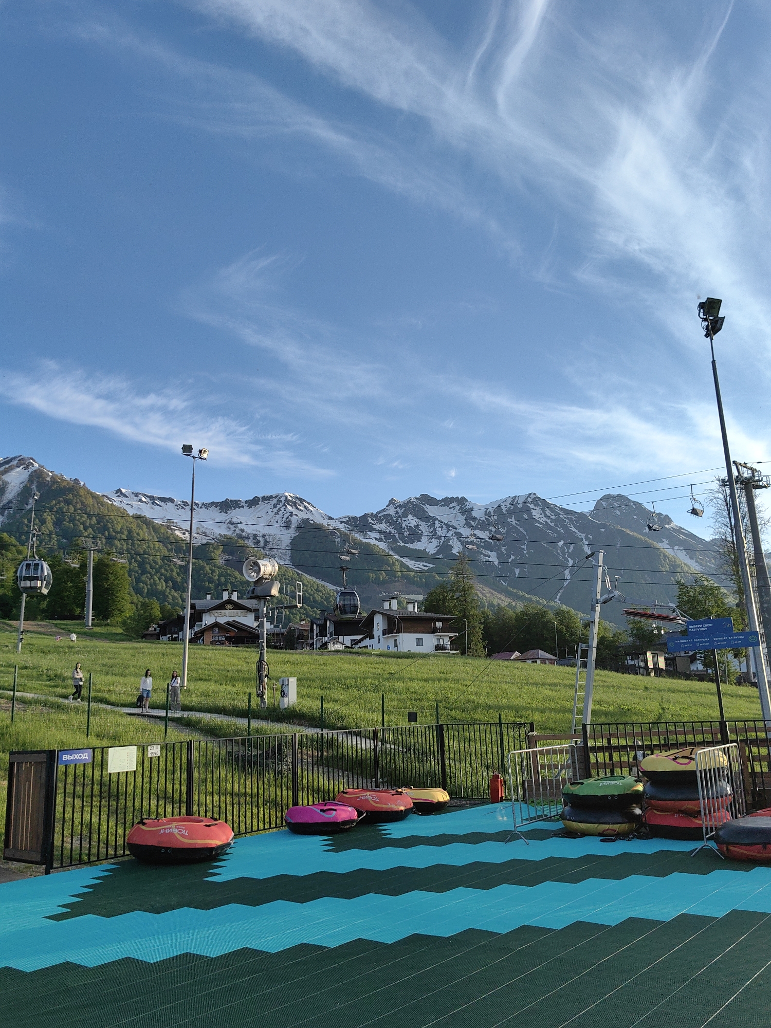 The image captures a vibrant, sunny day at a mountain resort, likely in late spring or summer. In the foreground, numerous inflatable tubing rings in red, orange, pink, green, yellow, and black are scattered and stacked on a distinct blue and dark green patterned mat. A black metal fence runs across the mid-ground, behind which a grassy slope rises. A wooden gate with the Cyrillic word "ВЫХОД" (Exit) is visible on the left, next to a small sign displaying numbers "23" and "1590".
Several people are walking on a paved path further up the grassy hill. In the middle ground, white resort buildings with dark roofs are nestled among lush green trees and slopes. One building prominently displays Cyrillic text, partially visible as "РОЗА...", suggesting a connection to the Rosa Khutor resort. Numerous ski lift poles, cables, and gondolas or chairlifts crisscross the scene, some with empty carriers. A large fan-like snowmaking device is also visible on a pole.
The background is dominated by majestic snow-capped mountains, showing clear peaks and forested lower slopes. The sky above is a brilliant blue with wispy, streaky white clouds. The overall lighting suggests it is daytime, possibly late afternoon, with clear weather. Another blue sign with white text is visible, indicating "ВЫБЕРИ СВОЮ ВАТРУШКУ" (Choose your tube) and offering options for "МАЛЕНЬКАЯ ВАТРУШКА" (Small tube) and "БОЛЬШАЯ ВАТРУШКА" (Large tube), implying dry tubing or summer fun activities at the resort. The text on the orange tubing rings appears to be "ЖОЕИНИ" or a similar brand name, visible in reverse.