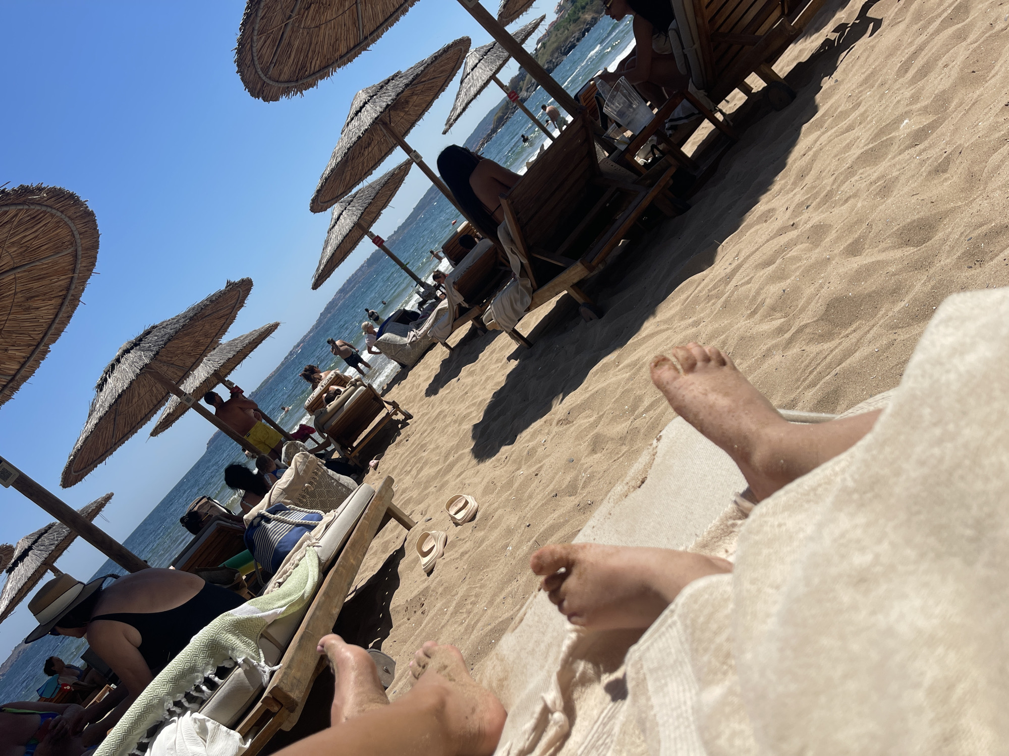 The image depicts a sunny day at a beach in Chernomorets, Bulgaria. Several individuals are visible, primarily relaxing on lounge chairs under thatched umbrellas. The foreground shows the bare feet of two individuals resting on a sandy surface, partially covered by a light-colored towel. In the mid-ground, people are seen sitting on lounge chairs, with some appearing to be reading or conversing. Further in the background, a man in yellow shorts stands near the water's edge, and other beachgoers are visible in the sea. The environment is characterized by golden sand, clear blue water, and a bright blue sky. The overall scene conveys a sense of leisure and vacation.