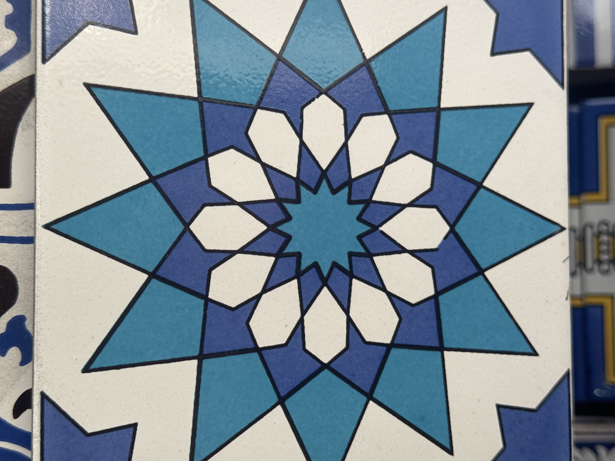 The image shows a close-up of a decorative tile with an intricate geometric pattern. The pattern consists of interlocking stars and polygons in shades of blue and white, outlined in black. The tile appears to be part of a larger tiled surface, as suggested by the edge visible at the top and bottom of the frame. There is no indication of the city of Niterói, Brazil, or any other specific location. The image focuses solely on the design of the tile.