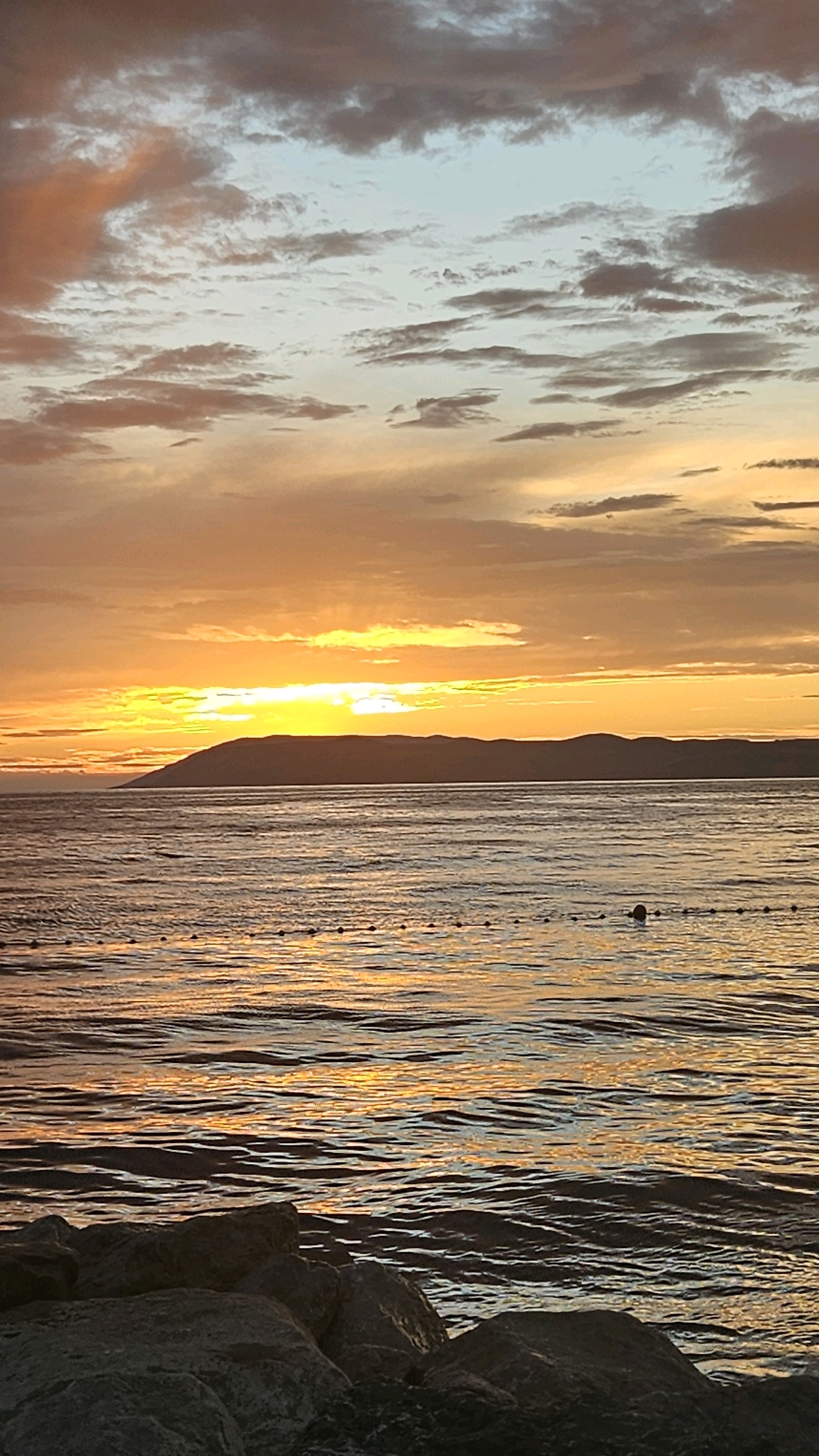 The image depicts a sunset over the sea in Općina Tučepi, Croatia. The sky is filled with dramatic clouds in hues of orange, yellow, and gray, illuminated by the setting sun. A prominent island or landmass with a mountainous silhouette is visible on the horizon. The sea in the foreground reflects the warm colors of the sky, with gentle ripples disturbing the surface. A line of buoys or markers stretches across the water, and a single dark object, possibly a buoy or a swimmer, is visible in the distance. In the immediate foreground, large, dark rocks are situated along the shoreline, suggesting a coastal or harbor area. The scene evokes a sense of tranquility and natural beauty.