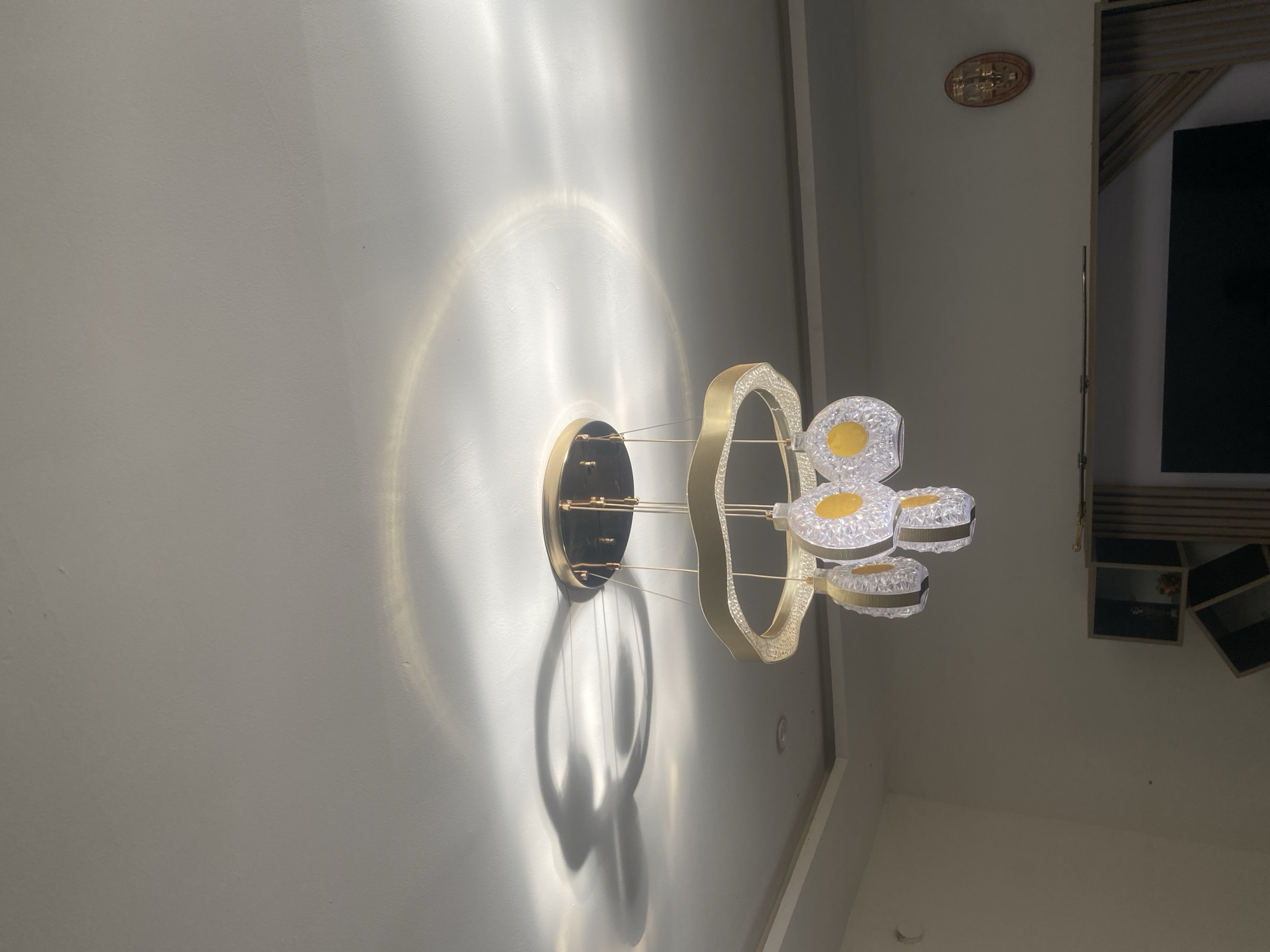 This media file presents an indoor scene, primarily showcasing a modern, wall-mounted light fixture. The fixture, centrally positioned, features a contemporary design with a prominent, irregular oval or ring-shaped structure in a gold-toned metallic finish, adorned with textured or faceted clear material that glows. Several smaller, circular or oval light elements, also with clear, textured surfaces and distinctive yellow centers, are attached to the main structure. These elements suggest integrated LED lighting. The entire fixture is mounted on a dark, circular base against a light-colored, subtly textured wall.
The light fixture is actively illuminated, casting a striking display of light and shadow. A large, soft halo of light emanates from behind the fixture's base, while intricate and sharp shadows, reflecting the fixture's complex geometric forms and thin connecting wires, are cast onto the wall below.
In the upper right corner of the frame, a small, oval-shaped decorative piece, possibly a traditional emblem or a clock, is affixed to the wall; however, any text or symbols on it are too indistinct to read. Below this, partially visible, are dark, modular shelving units or cabinetry and a large, dark rectangular screen, likely a television or monitor. The clean lines and modern design suggest a contemporary interior setting, such as a living area or corridor. The artificial lighting implies the scene takes place during evening hours or in an interior space where artificial illumination is consistently used. The location context is Abuja, Nigeria.