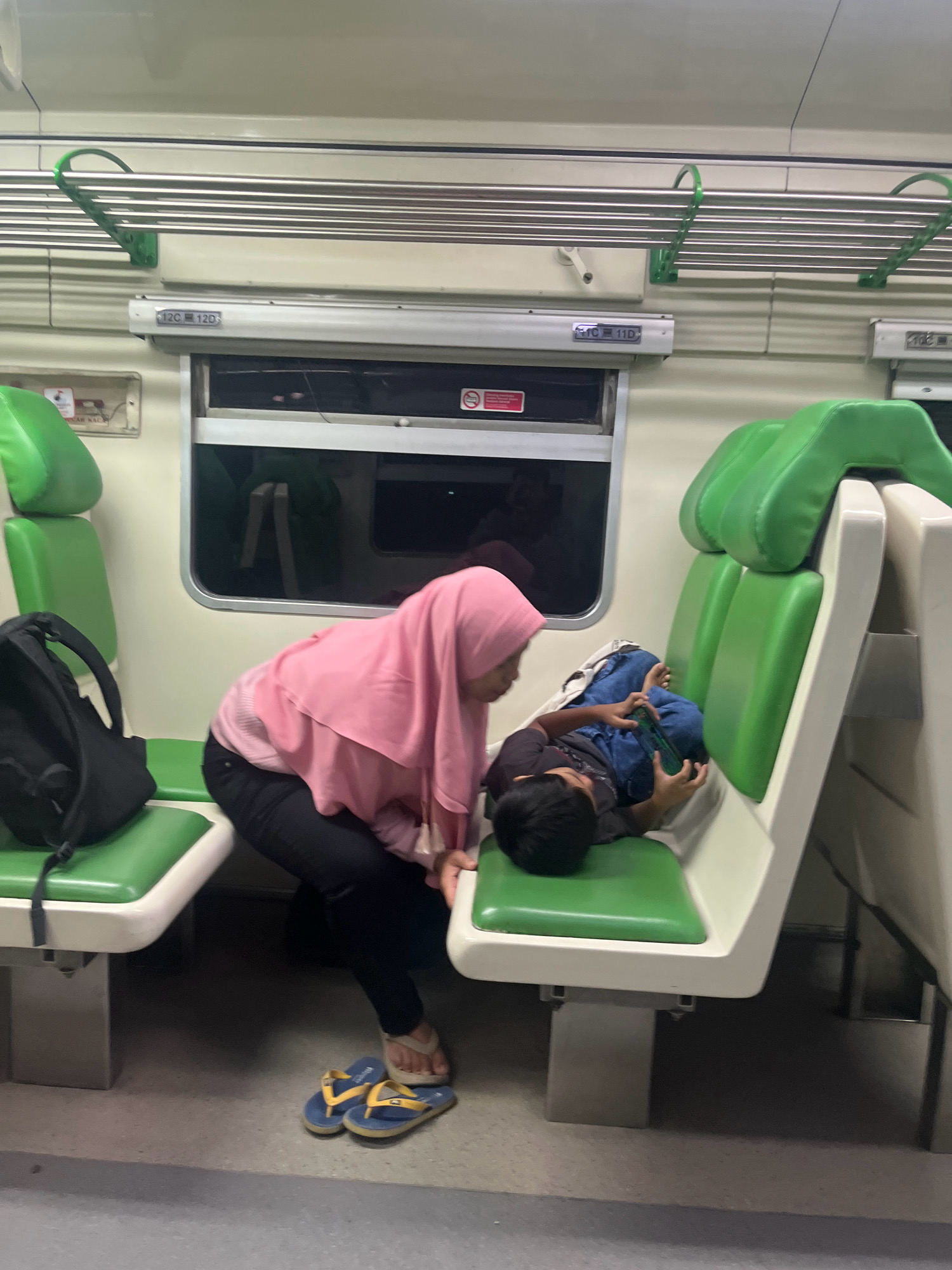 The image shows the interior of a train carriage, likely in Babat, Indonesia, based on the context provided. A woman wearing a pink hijab and black pants is tending to a young boy who is lying on a green seat, playing on a handheld video game. The seats are a vibrant green, and the carriage walls are a pale, neutral color. Above the window, a metal rack with several horizontal bars is visible. A black backpack rests on a seat to the left of the frame. The floor of the carriage is a muted gray.
