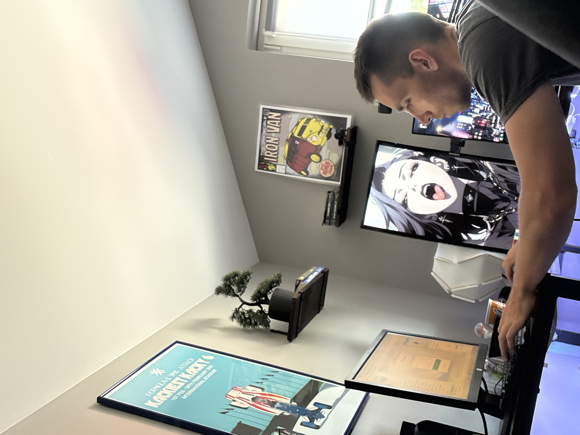 A man is shown from the side, looking at a computer monitor. He is wearing a gray t-shirt. The computer monitor in front of him displays an anime character. To his left, another monitor displays a webpage with text and a button. In the background, there are posters on the wall, including one with text that appears to read "KACKIEST KACKY 6." A small bonsai tree is on a shelf above one of the posters. The scene appears to be in a room with gray walls and a white ceiling. There is no indication of the location being in Gabrovo, Bulgaria.