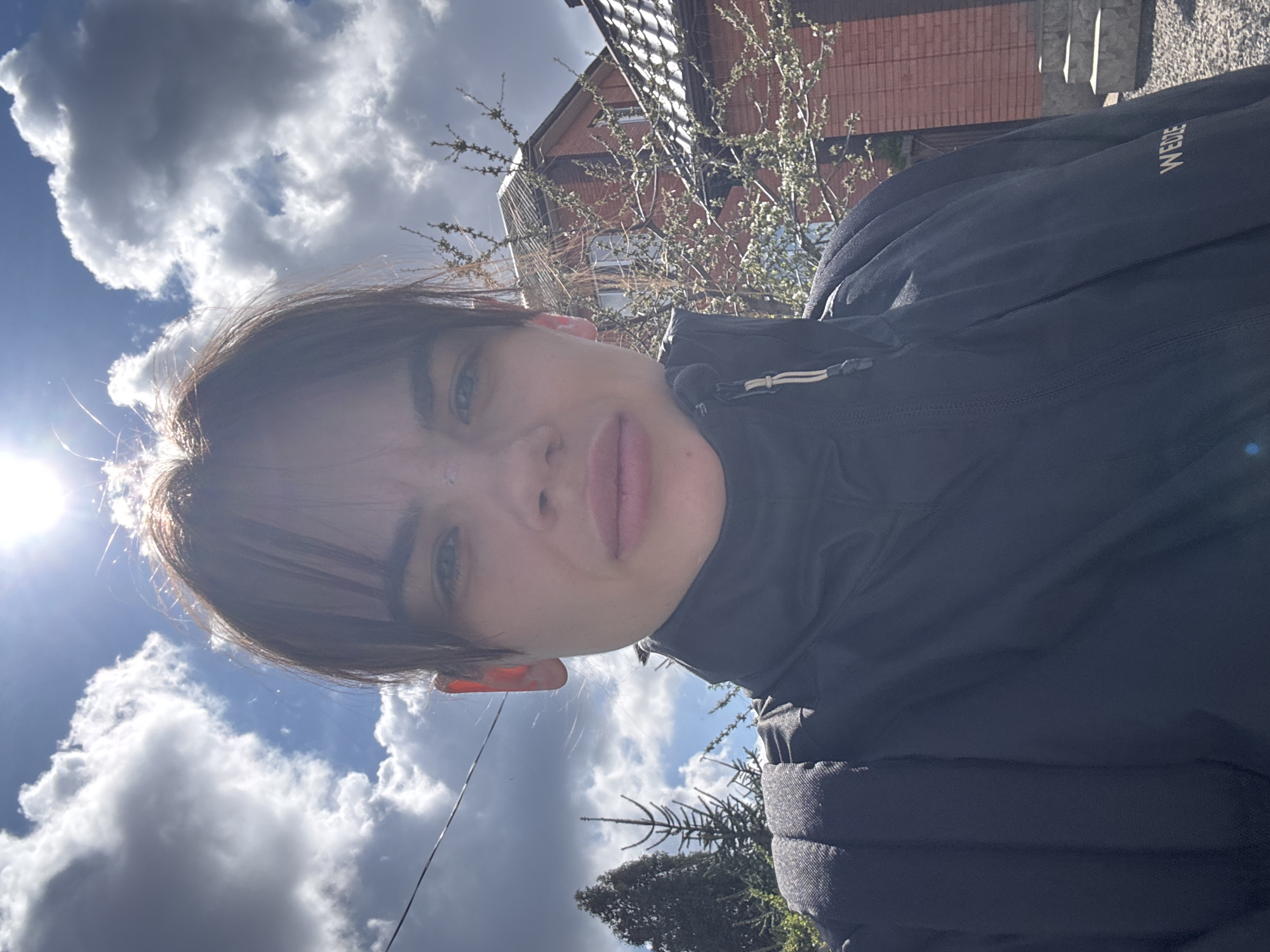 The image depicts a person wearing a dark-colored jacket with the brand name "WEDZE" visible. The photo is taken from a low angle, looking up at the person's face. The sky is partially cloudy with bright sunlight. In the background, there is a building, possibly a house, with a red roof and some trees. Power lines are also visible against the sky. The location is possibly in a residential area, but the specifics of Rostov-on-Don are unclear from this single image.