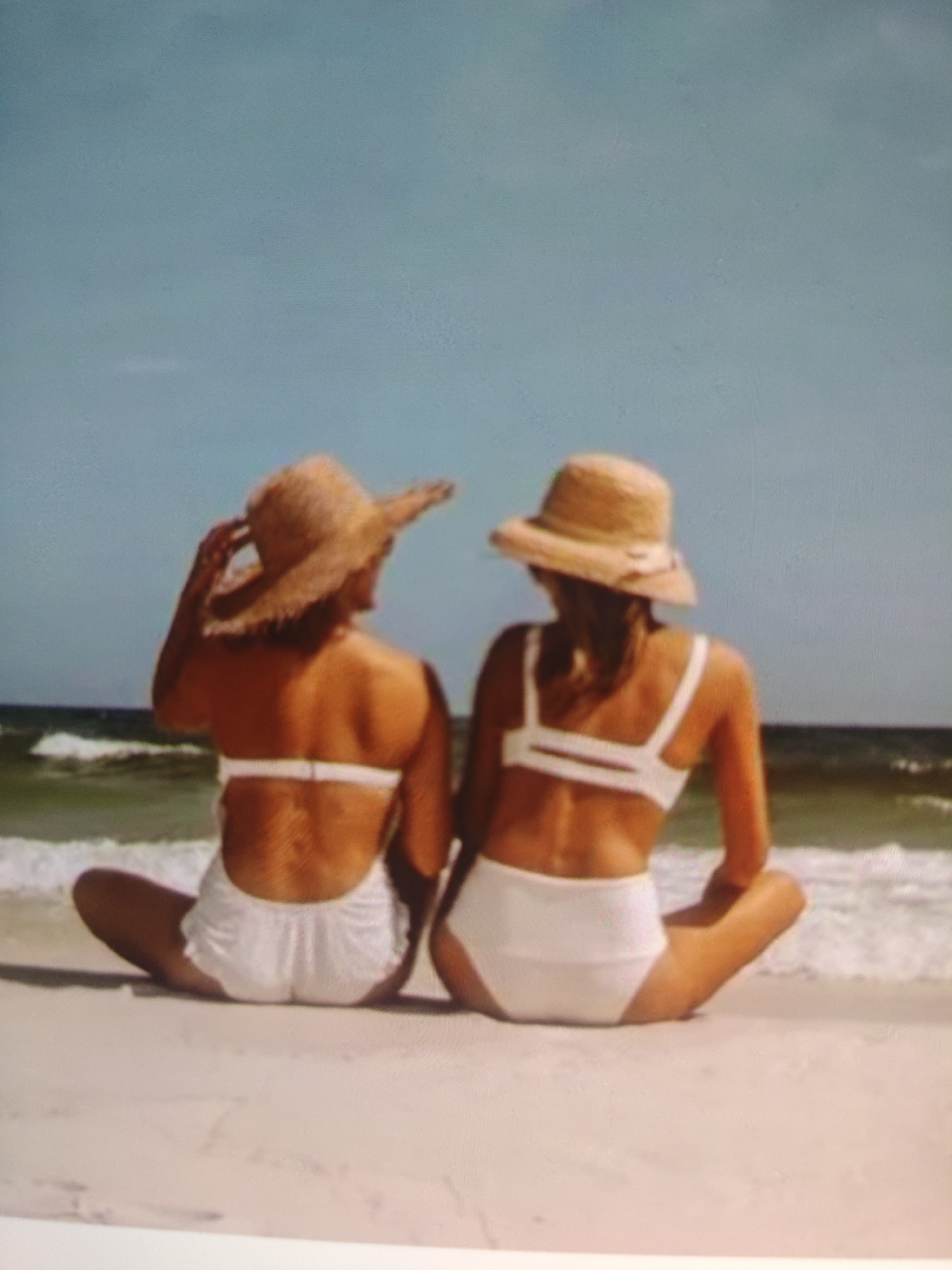 Two individuals, viewed from behind and sitting on a sandy beach, are depicted against a backdrop of the ocean and sky. Both are wearing straw hats and white bathing suits. The person on the left has their right hand on their hat, while the person on the right has their hands resting on their knees. The ocean has small waves breaking near the shore. The sky is a clear, light blue. The sand is a light tan color. The image is a photograph. This scene is not located in the city of Krasnaya Polyana, Russia.