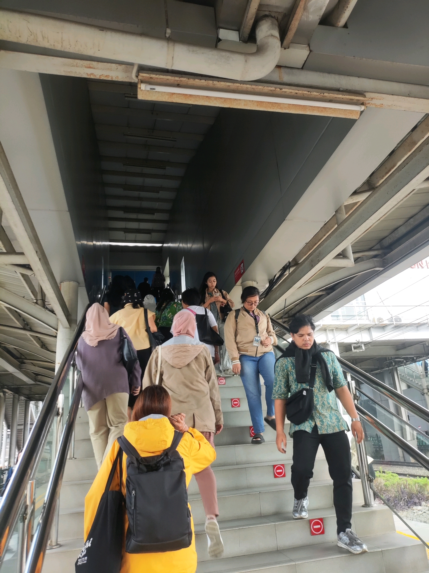 A group of people are walking up an outdoor staircase, likely an overpass or pedestrian bridge, in East Jakarta, Indonesia. The staircase is made of concrete with metal railings. There are "no standing" signs with a red circle and line on some of the steps. The environment appears urban, with visible infrastructure overhead, including pipes and lighting fixtures. The people are dressed in casual clothing, and some are carrying bags. The lighting suggests it is daytime.