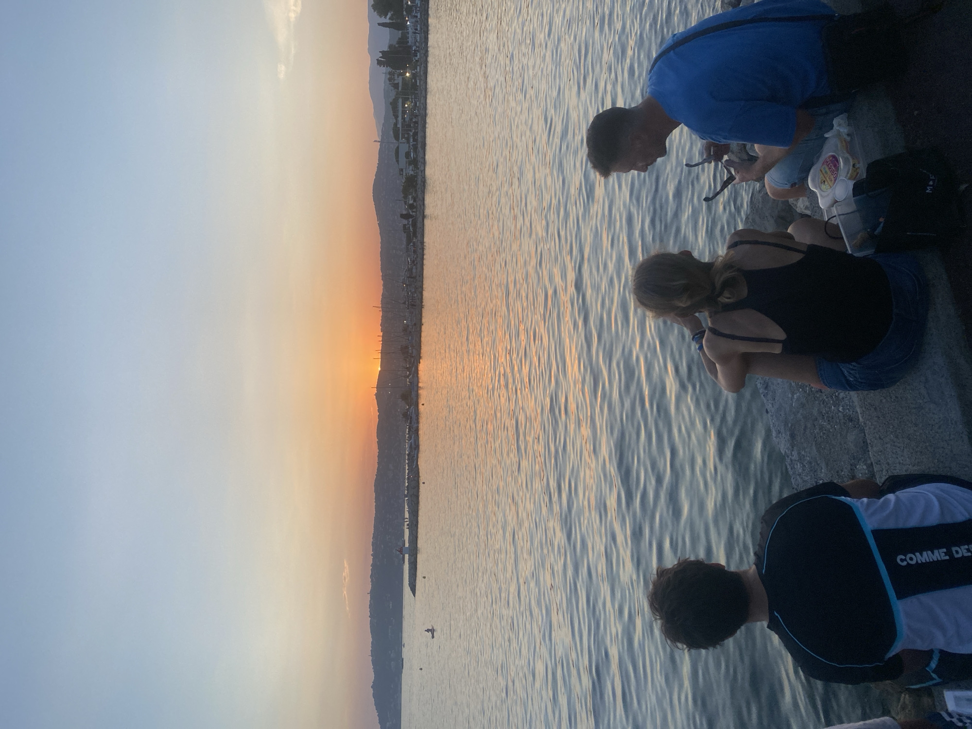 In Antibes, France, three people sit on a concrete seawall facing the Mediterranean Sea at sunset. The sky displays a gradient of colors from pale blue to orange and yellow as the sun dips below the horizon. The water is calm with gentle ripples reflecting the sky's hues.
On the left, a person with dark hair, viewed from behind, wears a black and white t-shirt with blue accents and the text "COMME DES" visible on the back. They are looking out at the sea.
In the center, a person with blonde hair, also seen from behind, wears a black tank top and denim shorts. They are looking towards the right of the frame.
On the right, a man with short brown hair, facing left, is wearing a bright blue t-shirt and light-colored shorts. He is holding a pair of sunglasses in his right hand and appears to be talking to the person in the center.
In front of them, on the ground, are a cooler bag, some food containers, and a black tote bag. In the distance, across the water, a peninsula is lined with buildings and trees. A lighthouse and a pier with people are visible, along with moored sailboats and a person on a paddleboard in the water. The overall atmosphere is serene and relaxed, characteristic of an evening by the sea.