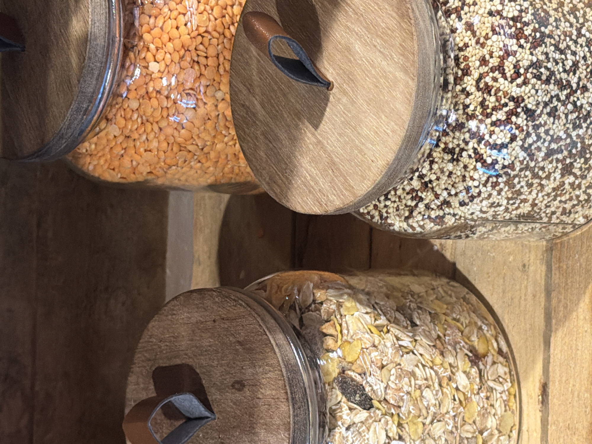 This is a close-up, low-angle shot of four glass jars filled with grains and legumes, displayed on a wooden shelf. The jars have round, wooden lids with small, dark grey straps attached to them. The grains are clearly visible through the glass. The top left jar contains bright orange lentils. Next to it, to the right, is a jar filled with tri-color quinoa, a mix of white, red, and brown seeds. The bottom jar, also on the left, contains a mixture of rolled oats and possibly other cereals. The contents of the fourth jar are mostly obscured, but appear to be similar to the quinoa. The scene is indoors, with warm, natural lighting that casts soft shadows, suggesting daytime. The wooden shelf and the overall aesthetic suggest a rustic or natural food store setting. There is no visible text in the image. The image is taken in Garmisch-Partenkirchen, Germany.