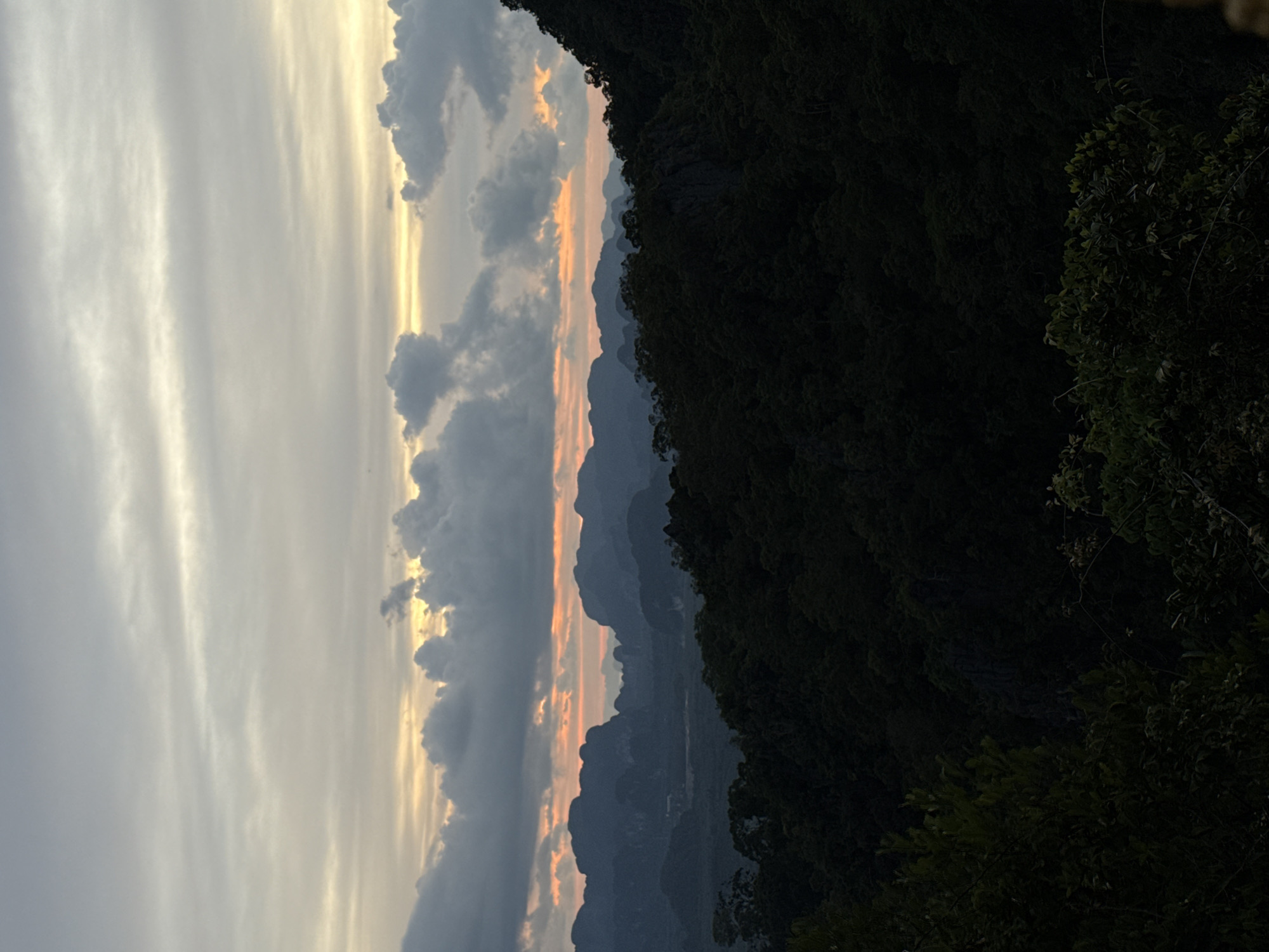 The image captures a panoramic view of a mountainous landscape at dusk. In the foreground, dense, dark green foliage of trees covers the lower portion of the frame, obscuring the ground. Beyond this, a series of silhouetted, jagged mountain peaks recede into the distance. The sky is a blend of soft grays and pale yellows, with streaks of orange and pink near the horizon, indicating the setting sun. Scattered, dark clouds drift across the sky, adding texture to the scene. The overall atmosphere is serene and natural, suggesting a remote or elevated vantage point, likely overlooking a valley or rural area within Ban Khao Tang, Thailand.