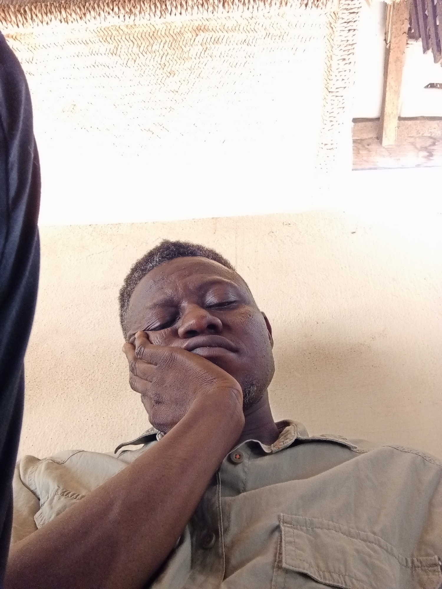 A person, with their eyes closed, rests their head on their hand. The person is wearing a light brown collared shirt with pockets. The background consists of a textured, light-colored wall and a woven ceiling. The scene appears to be indoors, possibly in a home or informal setting in Narayi, Nigeria.