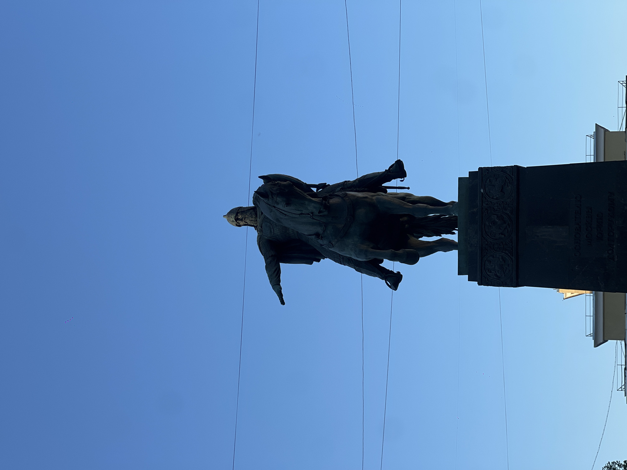 The image depicts the Monument to Yuri Dolgoruky, located in Tverskaya Square in Moscow, Russia. The monument features a large equestrian statue, cast in a dark material, likely bronze, silhouetted against a clear, bright blue sky.
The central figure is Yuri Dolgoruky, depicted as a bearded man wearing a helmet and armor. He sits atop a horse that appears to be in motion, with its front right leg slightly raised. The rider's left arm is bent, holding a shield close to his body, while his right arm is extended forward and slightly upward, with his hand pointing directly ahead. The horse's head is turned slightly to the right of the viewer's perspective.
The statue rests on a tall, dark, rectangular stone pedestal. The upper portion of the pedestal is adorned with relief carvings featuring intricate designs. Faint, dark Cyrillic text is visible on the lower face of the pedestal, although it is not fully legible.
The background consists of a uniformly bright, cloudless blue sky, occupying the majority of the upper frame. Several thin, dark horizontal lines, consistent with overhead power or tram wires, traverse the sky at various heights, crossing in front of and behind parts of the statue. In the far distance, several small dark specks, possibly birds or distant objects, are scattered across the sky. The statue is primarily backlit, casting it into a strong silhouette against the bright sky. Below the pedestal, indistinct architectural elements of distant buildings are visible, suggesting an urban environment.