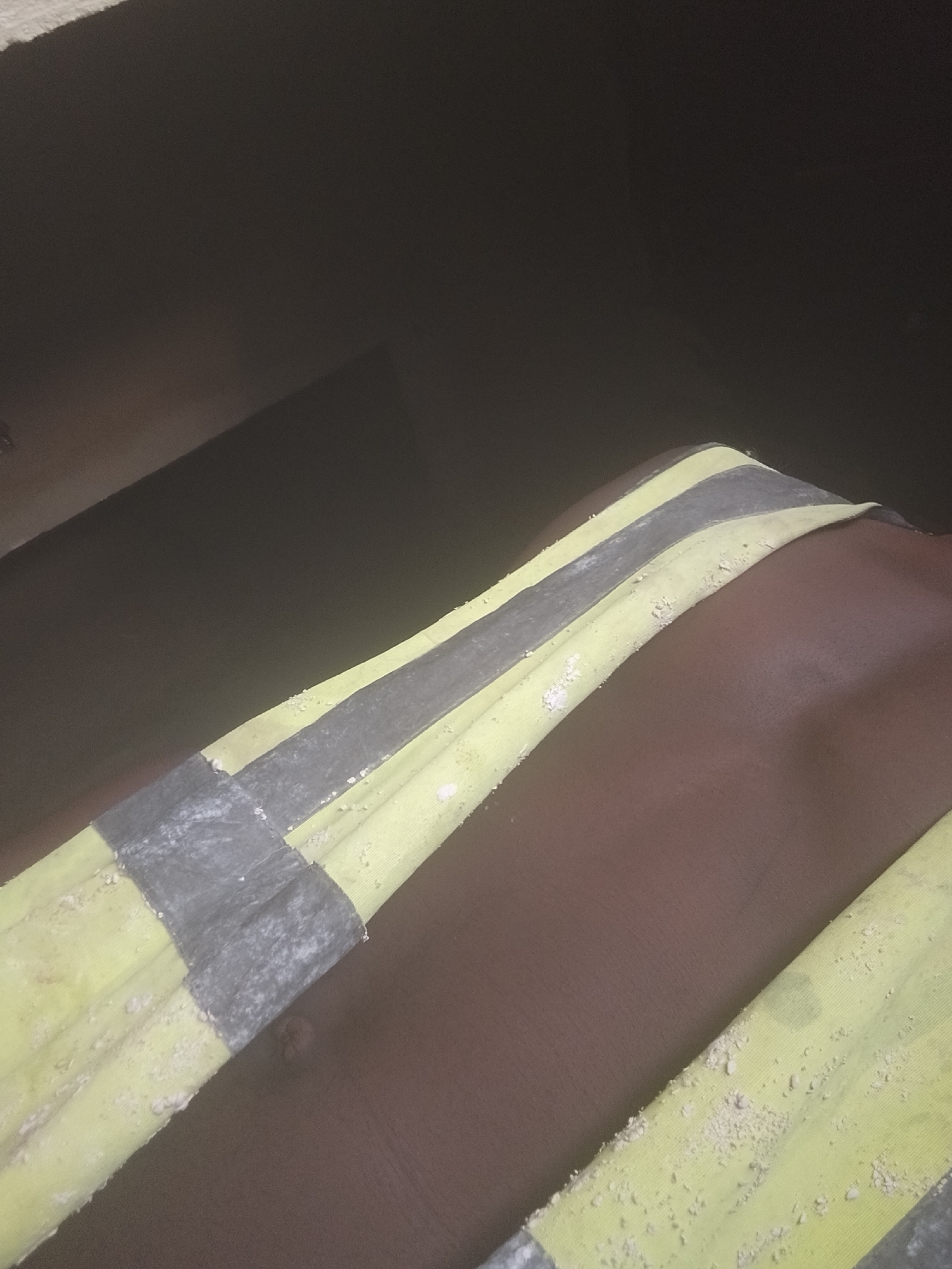 The image shows a close-up of a person's upper body, with a reflective vest draped over their torso. The vest is yellow with grey reflective stripes and appears to be slightly worn, with some dust or debris on its surface. The person's skin is dark brown and smooth, with a slight sheen. The background is dark and out of focus, suggesting a dimly lit environment. No specific location in the city of Unknown, Unknown can be determined from the image.
