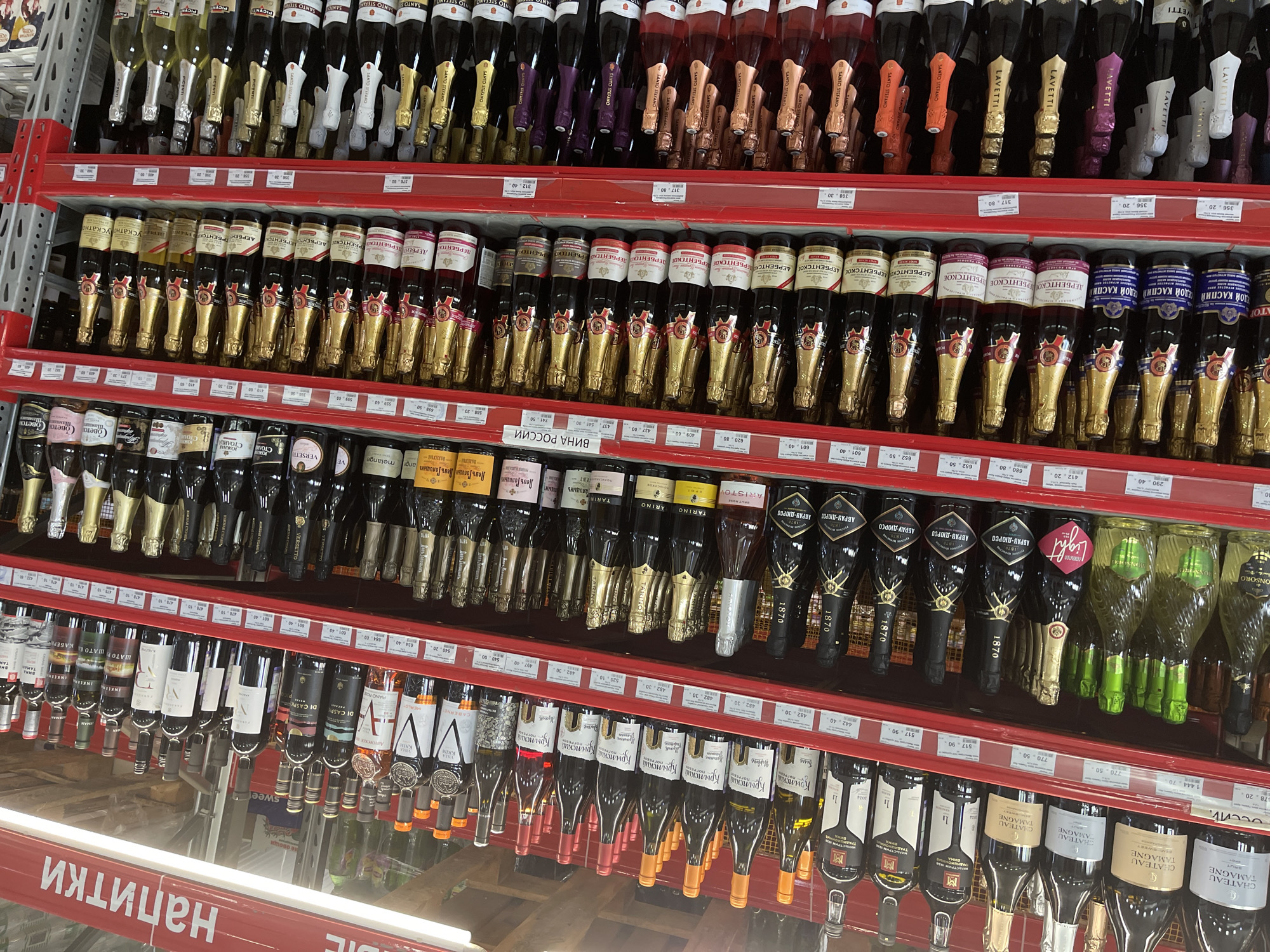 The image displays a retail environment, likely a supermarket or liquor store, stocking a wide variety of alcoholic beverages, primarily wine and sparkling wine. The shelves are organized with multiple rows of bottles, most of which are glass and have prominent labels indicating brand and type of beverage. Many of the bottles are in green, clear, and dark glass, with a significant portion being sparkling wine bottles in gold and silver foil wrappers. The overall color scheme is dominated by red shelving units and the varied colors of the bottle labels and packaging.
The location is identified as Aleksandrovskoye, Russia, suggested by the Russian text visible on some of the labels, such as "Напитки" (Drinks) and other Cyrillic script on the wine bottles. The lighting appears to be artificial, typical of an indoor retail setting. There are no people visible in the frame. The image focuses on the products for sale, with a strong emphasis on the presentation of these beverages on the shelving. The composition is that of a product catalog shot or a shelf view from a customer's perspective.