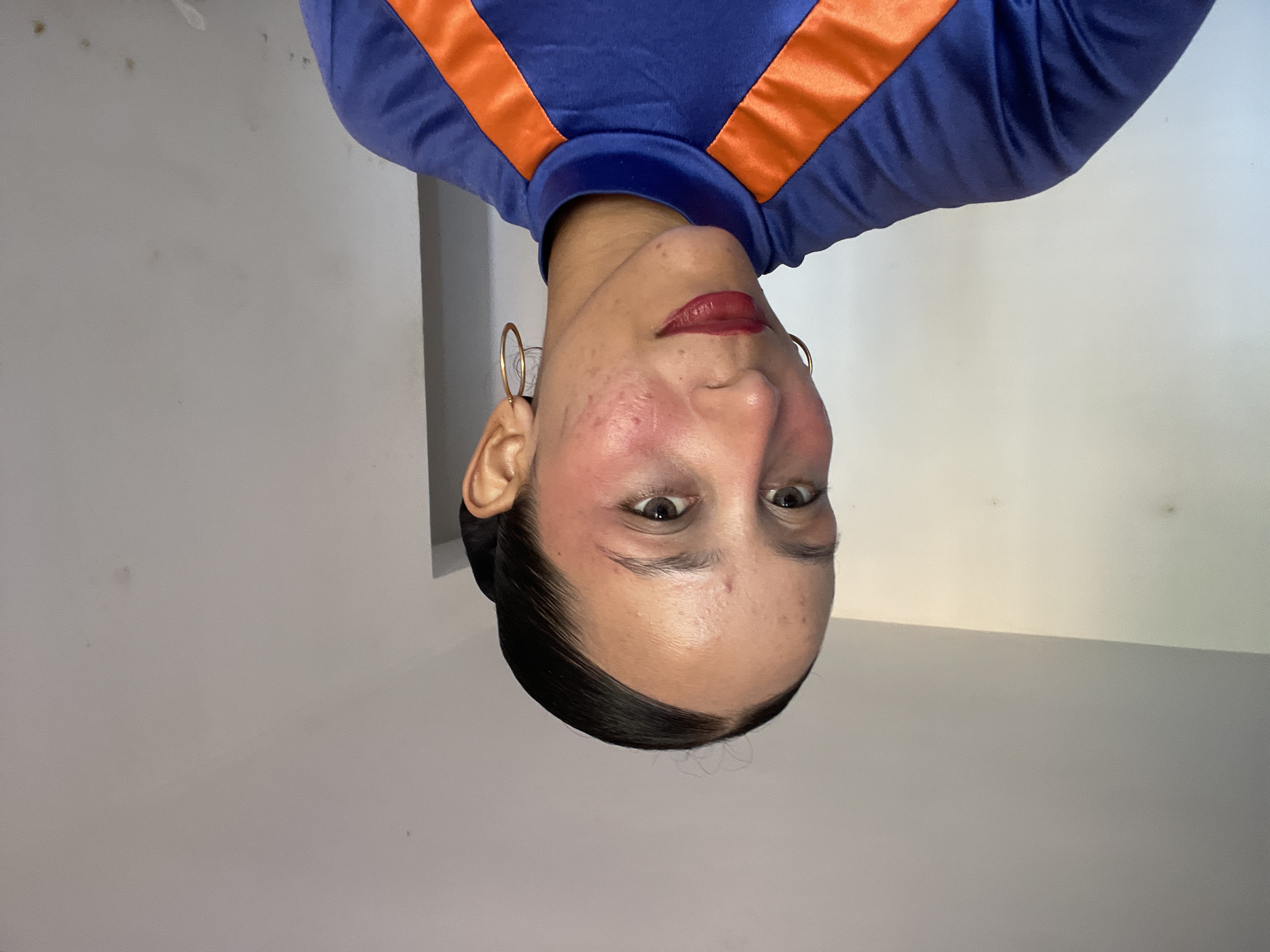 A medium shot shows a young woman with dark hair pulled back into a bun, wearing a blue and orange top. She is looking slightly to her left, with her head tilted down. Her makeup includes red lipstick and blush applied to her cheeks and temples. She has hoop earrings on both ears. The background is a plain, light-colored wall with some subtle texture. The lighting appears to be natural, coming from the left side of the frame. There is no visible indication of the city of Porlamar, Venezuela.