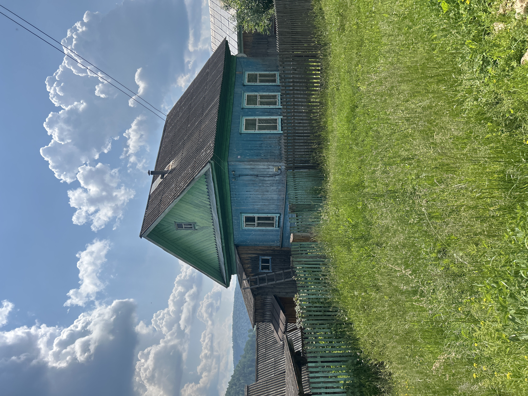 In Новомуллакаево, Russia, a traditional wooden house painted bright blue with green trim stands under a cloudy blue sky. The house has a dark, weathered roof and multiple windows with light-colored frames. A green wooden fence encloses the property, which is surrounded by tall, green grass and wildflowers. Adjacent to the main house, there is a smaller wooden structure with a sloped roof and a ladder leaning against it. In the background, a hilly, tree-covered landscape is visible, and power lines cross the sky above. The scene appears to be a rural residential setting.