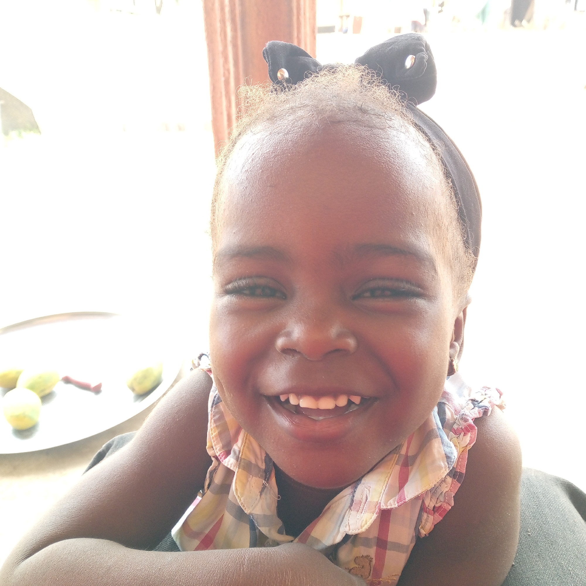 A close-up, brightly lit shot shows a young, dark-skinned girl with a beaming smile, wearing a black headband with decorative bows. She has short, curly hair and wears a collared, plaid shirt. Her arms are crossed in front of her. To the left of her is a silver platter with several yellow-green fruits and a red object on it. The background is blurred and appears to be indoors, though visibility is limited. There is no contextual information to confirm a location in Mararaba, Nigeria.