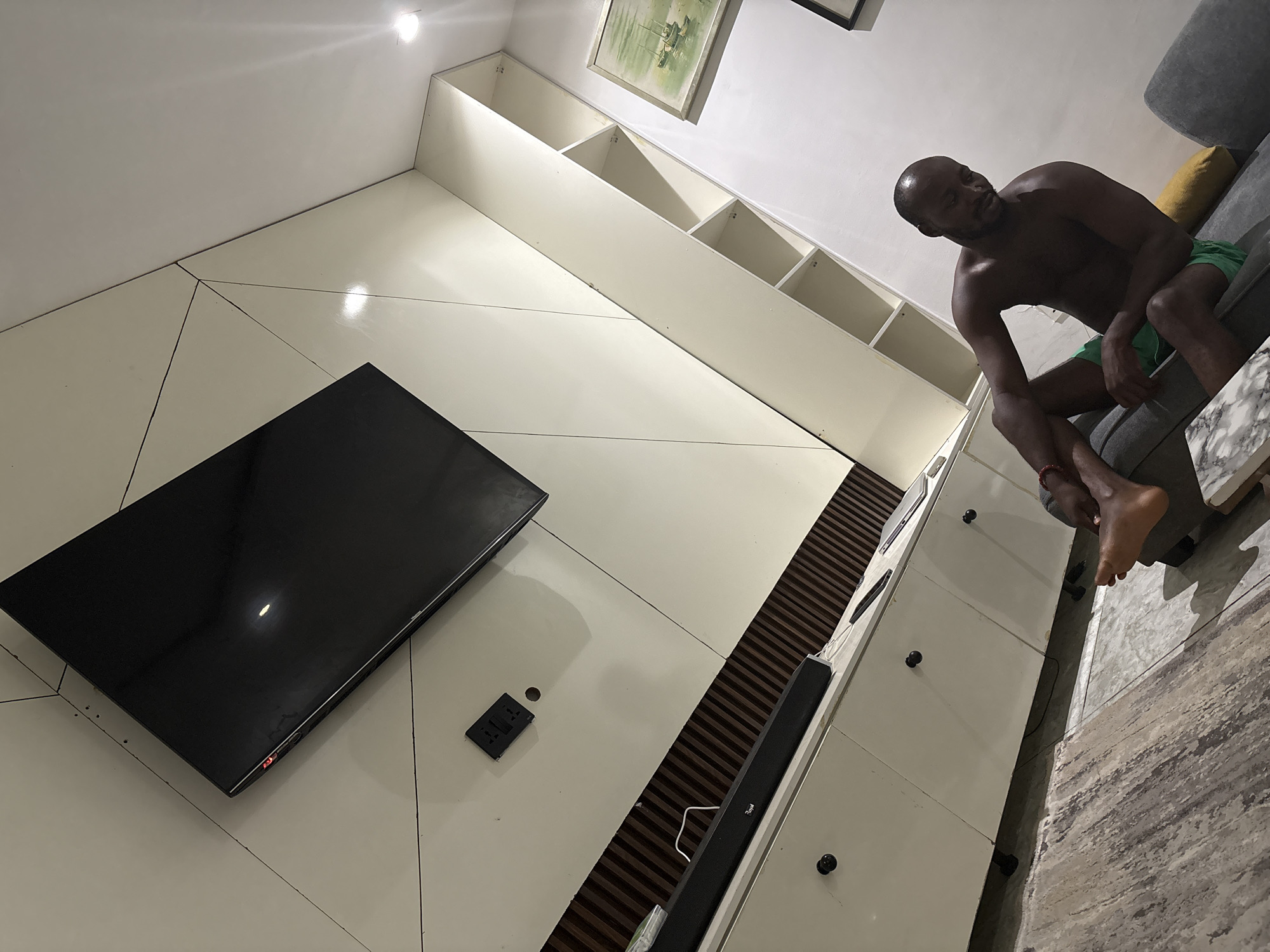 A man is sitting on a grey couch in a room with a white wall-mounted entertainment center. The TV is black and wall-mounted on the left. A soundbar and various shelves and cabinets make up the entertainment center. The flooring is a light grey textured material. The location is unknown.