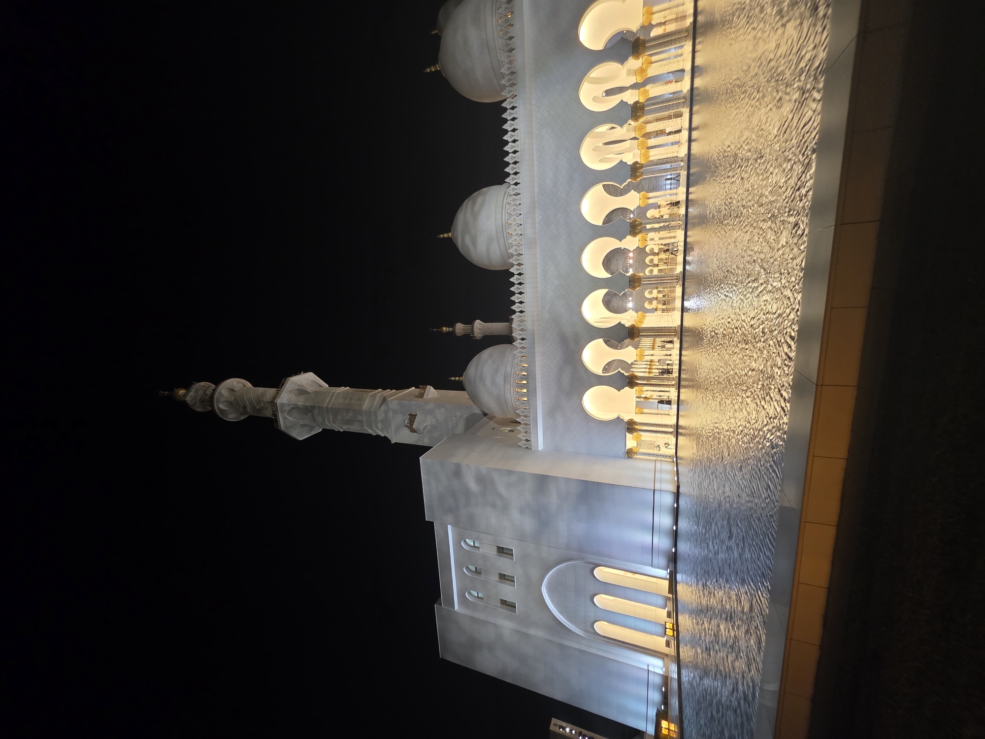 The image shows the Sheikh Zayed Grand Mosque in Abu Dhabi, United Arab Emirates, at night. The mosque is illuminated by white and yellow lights, creating a striking contrast against the dark sky. A large reflecting pool in the foreground mirrors the lights and the architecture of the mosque. The main building features white marble walls, multiple domes, and a tall minaret. Arches with decorative columns line the perimeter of the mosque. There are a few indistinct figures visible in the distance walking along the illuminated arcade. The overall scene conveys a sense of grandeur and tranquility.