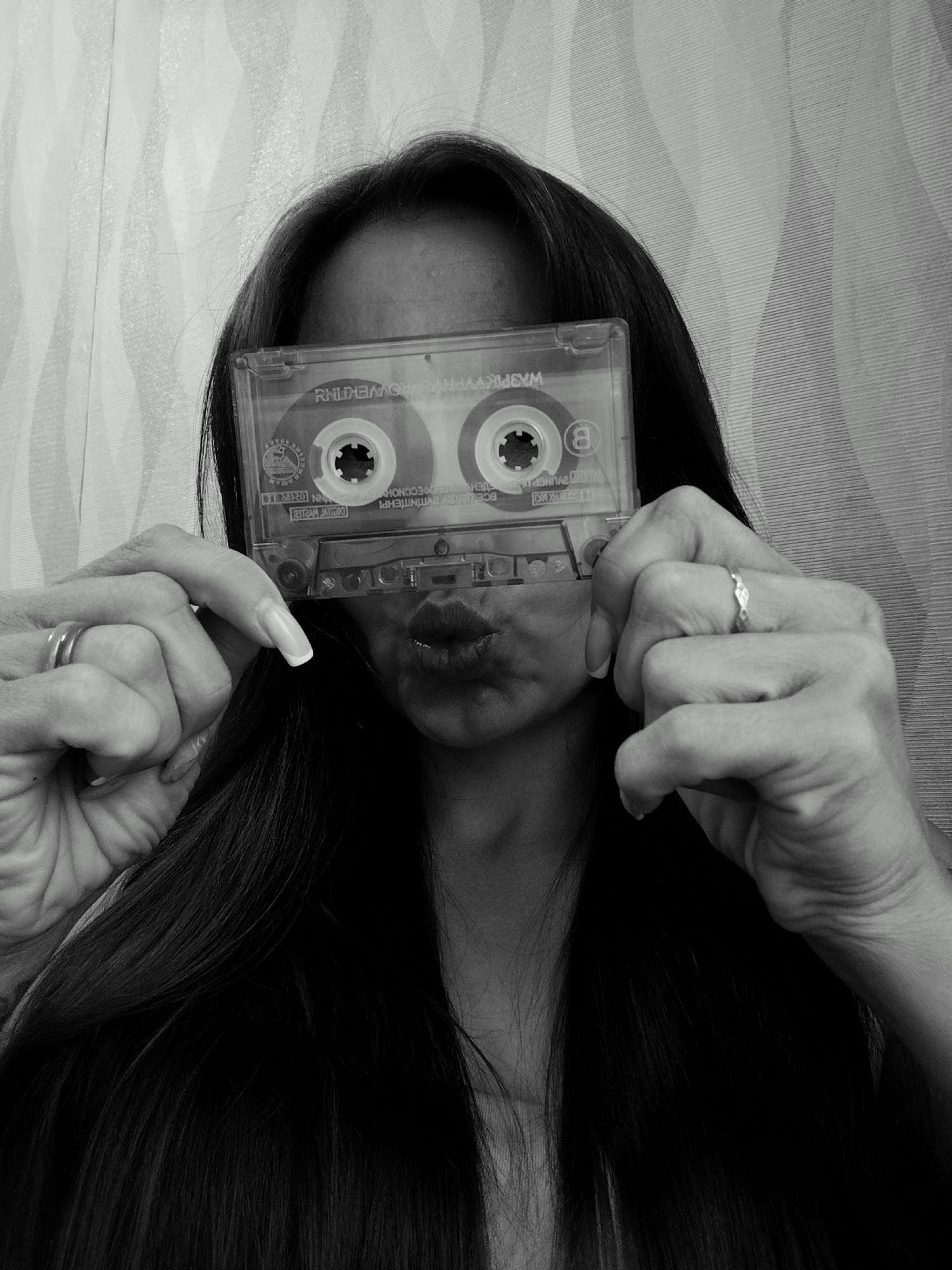A woman with long, dark hair is holding a clear cassette tape in front of her face, obscuring her nose and eyes. Her lips are visible through the tape, puckered in a kiss or pout. Both hands are visible, each with a ring on one of the fingers. The background is a neutral-toned wall with a subtle patterned design. The image is in black and white. The location of Фоки, Russia is not identifiable in the image.