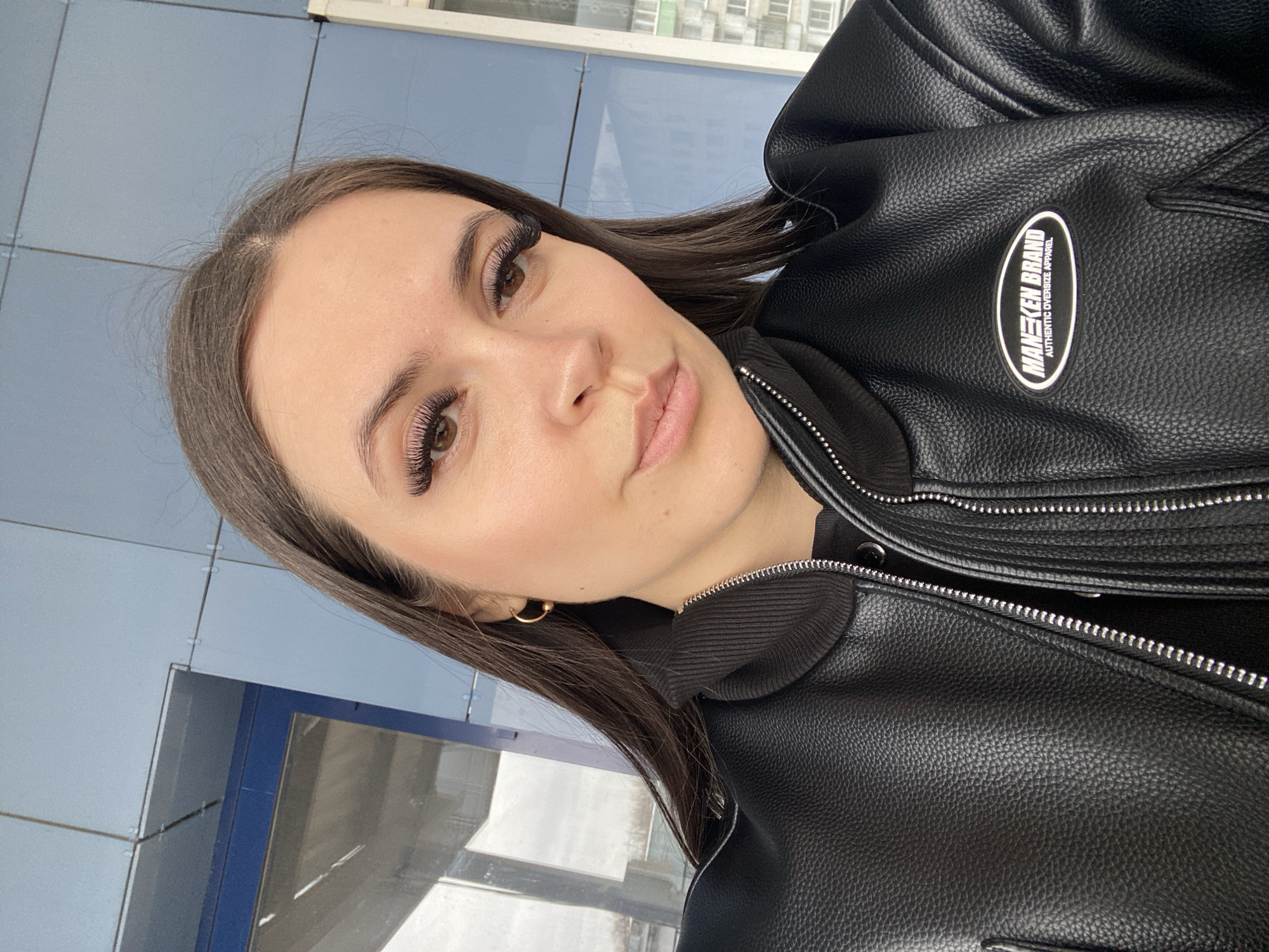 A medium shot photograph depicts a light-skinned woman with long, dark hair and false eyelashes wearing a black leather jacket. The jacket has a visible silver zipper and a logo on the right side that reads "MANKEN BRAND, AUTHENTIC OVERSIZE APPAREL." The woman is wearing a small gold hoop earring in her left ear.
The background suggests an urban setting with light blue metal paneling, likely a building facade. Part of a window or reflective surface is visible, showing a glimpse of what appears to be another building with windows. The scene appears to be located in Chaikovsky, Russia.