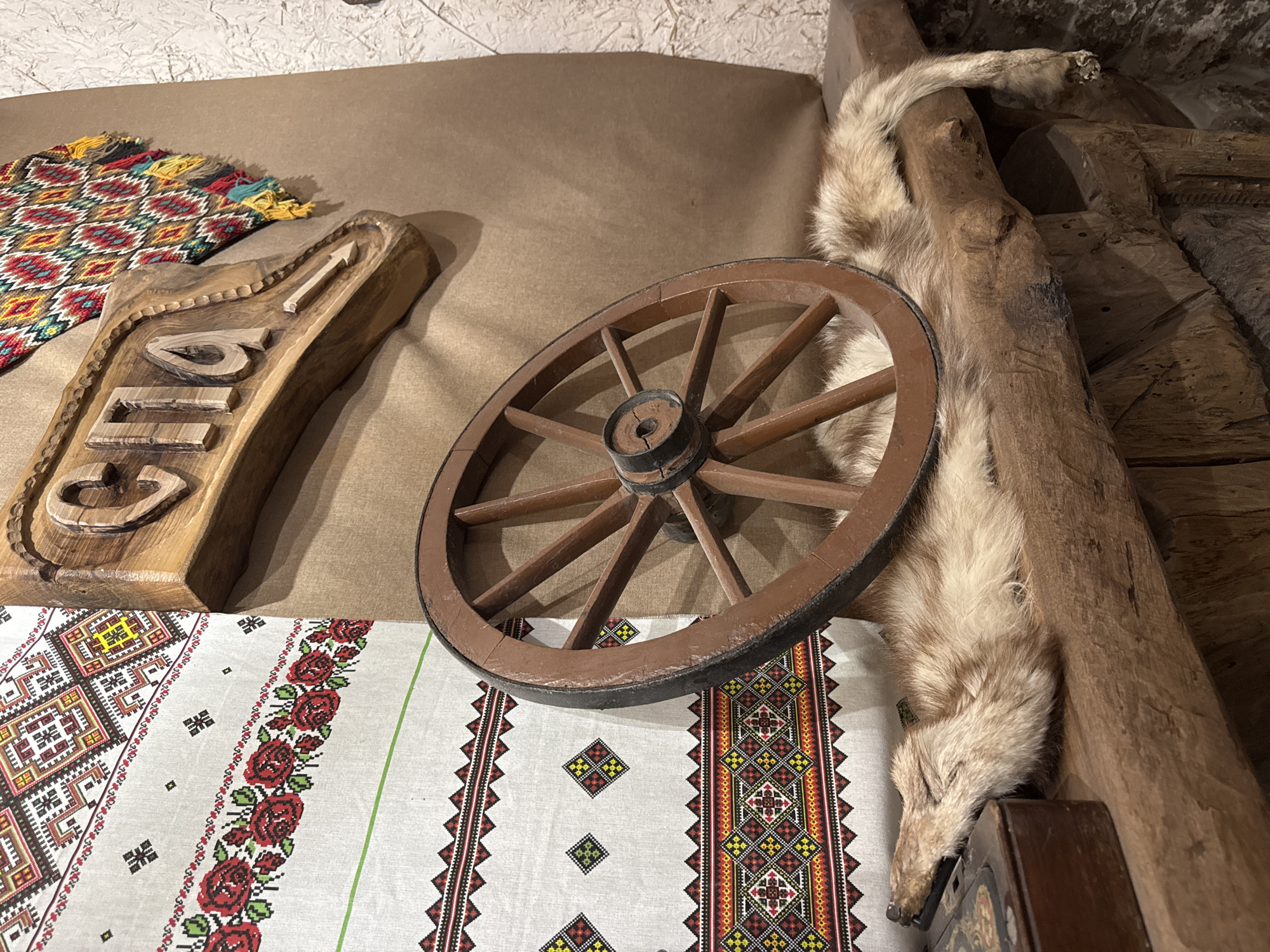 The image captures a rustic interior scene, likely an ethnographic display. A wooden wagon wheel leans against a fur pelt and a rough-hewn wooden post. A carved wooden sign is visible, partially obscured by a hand-woven textile with geometric patterns. Another colorful woven textile with fringes lies to the left of the sign. The scene is illuminated with natural light. Location unknown other than the context provided. The display appears to showcase traditional crafts and materials.