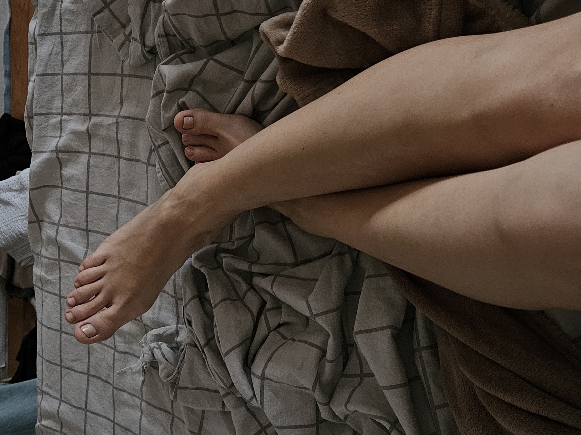 A close-up, elevated shot depicts a person's crossed legs and feet resting on a gray and white checkered bedding with a brown blanket partially covering the right leg. The toenails are painted with a neutral color. The background suggests an interior setting. The location of the city of Chaikovsky, Russia, cannot be confirmed based solely on visual information.