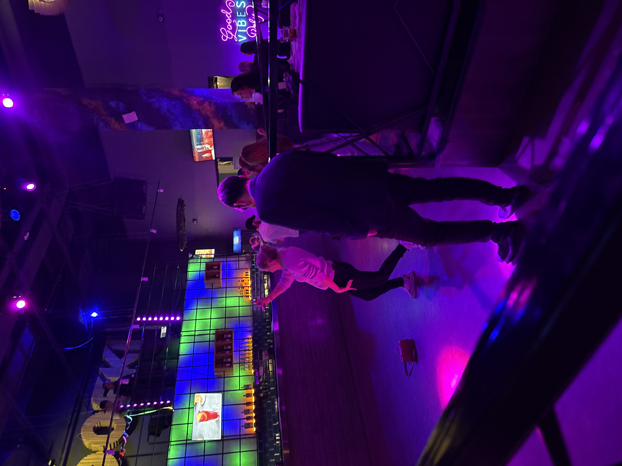 The image shows the interior of a bar or restaurant in Perm, Russia. The walls are decorated with bright, neon-lit patterns, including blue and green squares and a "Good Vibes Only" sign. Several people are visible, including a man in a dark jacket standing near the center of the frame and a woman in a light-colored shirt reaching towards the colorful wall display. A small brown purse rests on the floor nearby. The overall lighting is dark and dominated by purple hues.