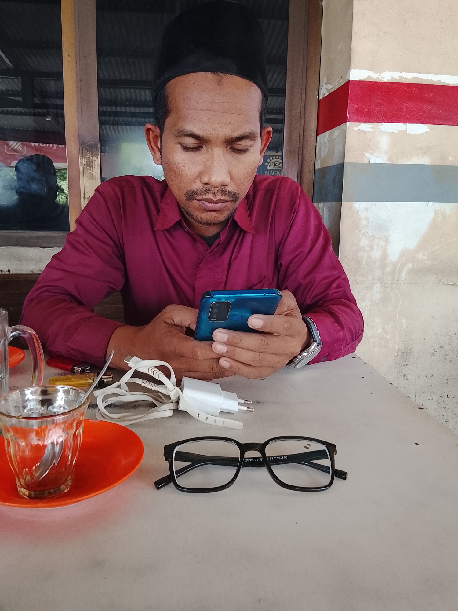 A medium shot captures a man wearing a maroon collared shirt and a black peci cap, seated at a table in what appears to be a roadside cafe in Lhokseumawe, Indonesia. He is looking down at a blue smartphone he holds in his hands, with a focused expression. On the table in front of him, a clear glass with a spoon sits on an orange saucer. Nearby, there are two lighters, a white charging cable, and a white USB adapter. To the right of these items, a pair of black-rimmed glasses rests on the table. The background shows a window with a reflection of another person, and a wall painted with red and grey stripes. The overall setting suggests a casual, everyday scene in a public eatery.