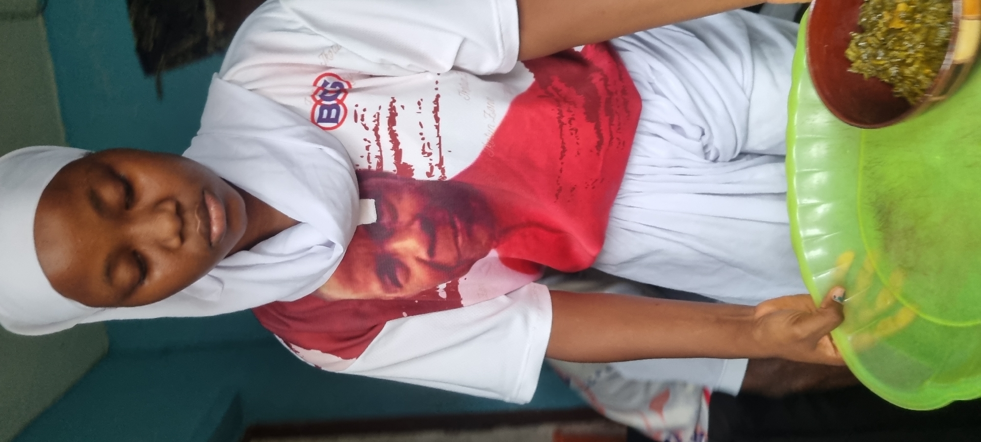 In Egan, Nigeria, a young woman wearing a white hijab and a t-shirt with a picture of an older man's face on it is holding a green plastic tray with a brown bowl of leafy green food on the right side of the tray. She is looking down at the tray. The background appears to be a room with a blue wall and a window.