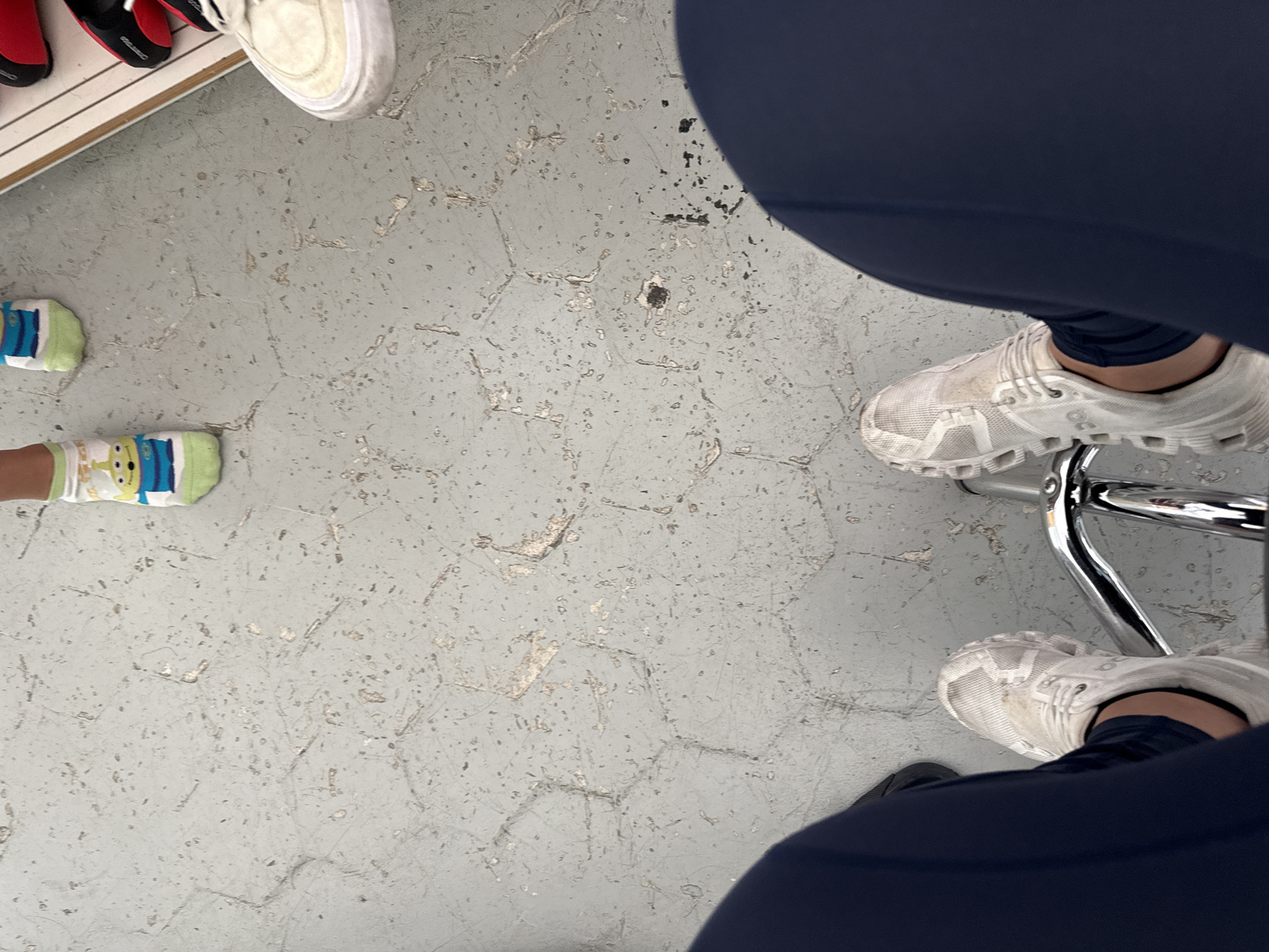 The image shows a gray, hexagonal-tiled floor. Near the top-left corner is a shoe rack with shoes. To the left of the center is a child's pair of socks with cartoon alien designs. At the right are two pairs of white athletic shoes, partially obscured by dark-blue athletic pants. There is a metal chair leg near the top-right. The location is not identifiable from this image.