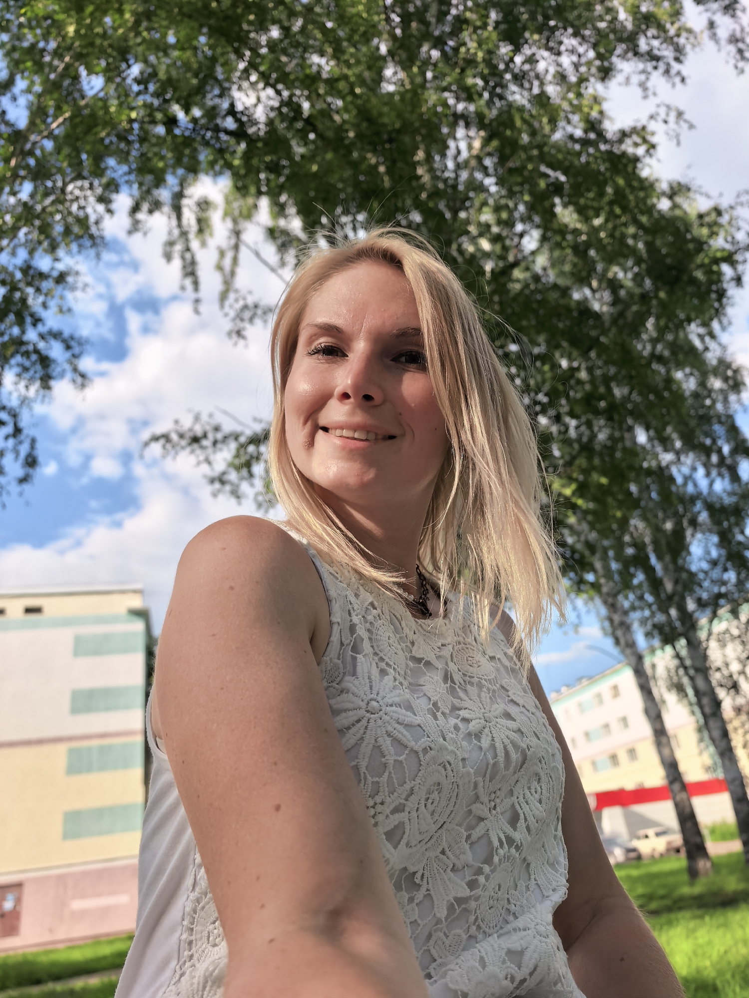 This media file is a selfie taken outdoors in Sterlitamak, Russia. The subject is a young woman with blonde hair, smiling and looking at the camera. She is wearing a white lace top. Behind her, the background consists of green trees with dappled sunlight filtering through, and a blue sky with white clouds. In the distance, there are buildings with light-colored facades and colorful accents, suggesting an urban or suburban environment. The overall mood is cheerful and relaxed, possibly indicating a sunny day. The time of day appears to be daytime, likely late morning or afternoon, given the brightness of the light. There is no visible text in the image.
