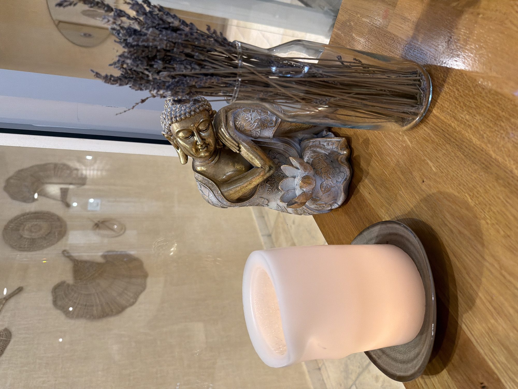 In the city of Galil Elyon Regional Council, Israel, a close-up, eye-level shot captures a tabletop vignette. On the left, a large, white, unlit LED candle rests on a dark grey, irregularly shaped ceramic coaster. To the right of the candle, a small, golden Buddha statue is positioned, with its head resting on its arm. The statue is adorned with intricate patterns and a lotus flower at its base. Beside the Buddha, a clear glass vase holds a bunch of dried lavender. The objects are arranged on a warm-toned wooden table, reflecting light. In the background, a wall with decorative woven circular art pieces and a partial view of a window are visible. The lighting is soft, creating a serene atmosphere.