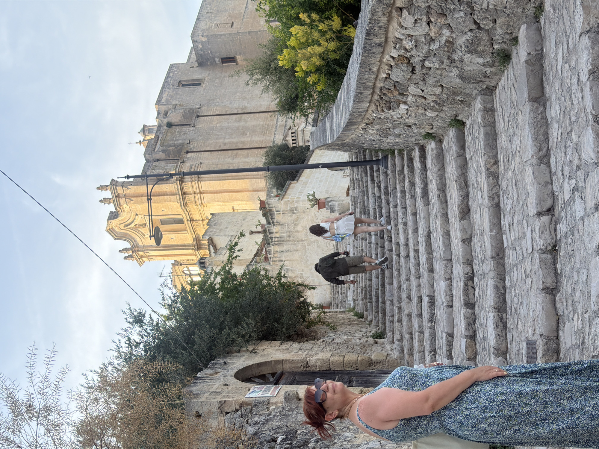 In Matera, Italy, on a sun-drenched afternoon, a woman with auburn hair pulled back and wearing large sunglasses and a blue floral dress stands on the left side of a stone staircase. She is looking towards the right, her body angled slightly away from the camera. Her left hand rests on her hip, and she wears a gold ring on her right hand. To her right, two individuals are ascending the stone steps. The person in front, wearing a dark olive-green jacket and grey shorts, is walking uphill. Behind them, a person in a white crop top and white skirt is following.
The staircase is constructed from large, weathered stone blocks, worn smooth by time and foot traffic. A thick, rough-hewn stone wall runs along the right side of the stairs, with sparse green vegetation growing between the stones. Overhead, the branches of a dry, leafless tree extend from the left side of the frame. Further up the stairs and in the background, a large, ancient building made of pale stone dominates the scene. Its facade features classical architectural elements, including a prominent, ornate pediment at the top. A street lamp with a dark fixture is positioned on the left side of the steps, near the building. The sky above is a clear, pale blue with a few wispy clouds.